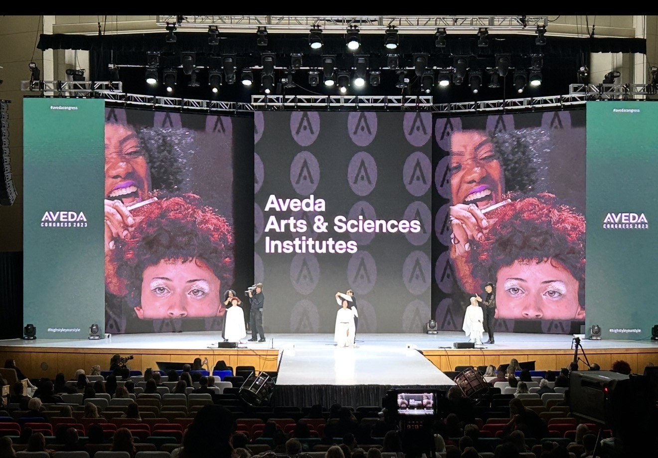 Stage with large screens displaying smiling faces and text 'Aveda Arts & Sciences Institutes', audience seated.
