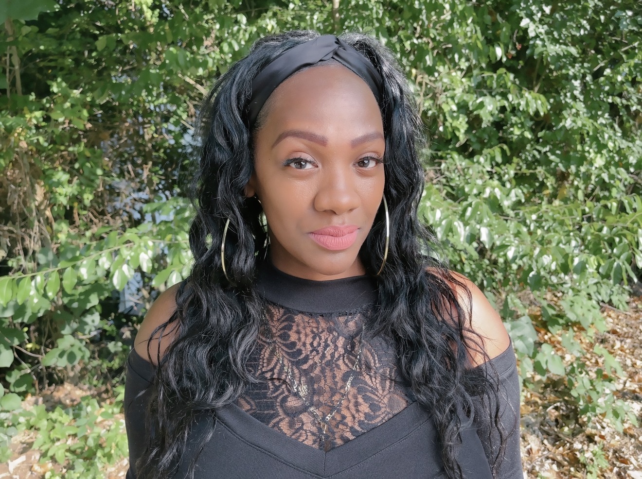 Woman with long wavy hair and hoop earrings outdoors, wearing a black lace top, standing in front of green foliage.