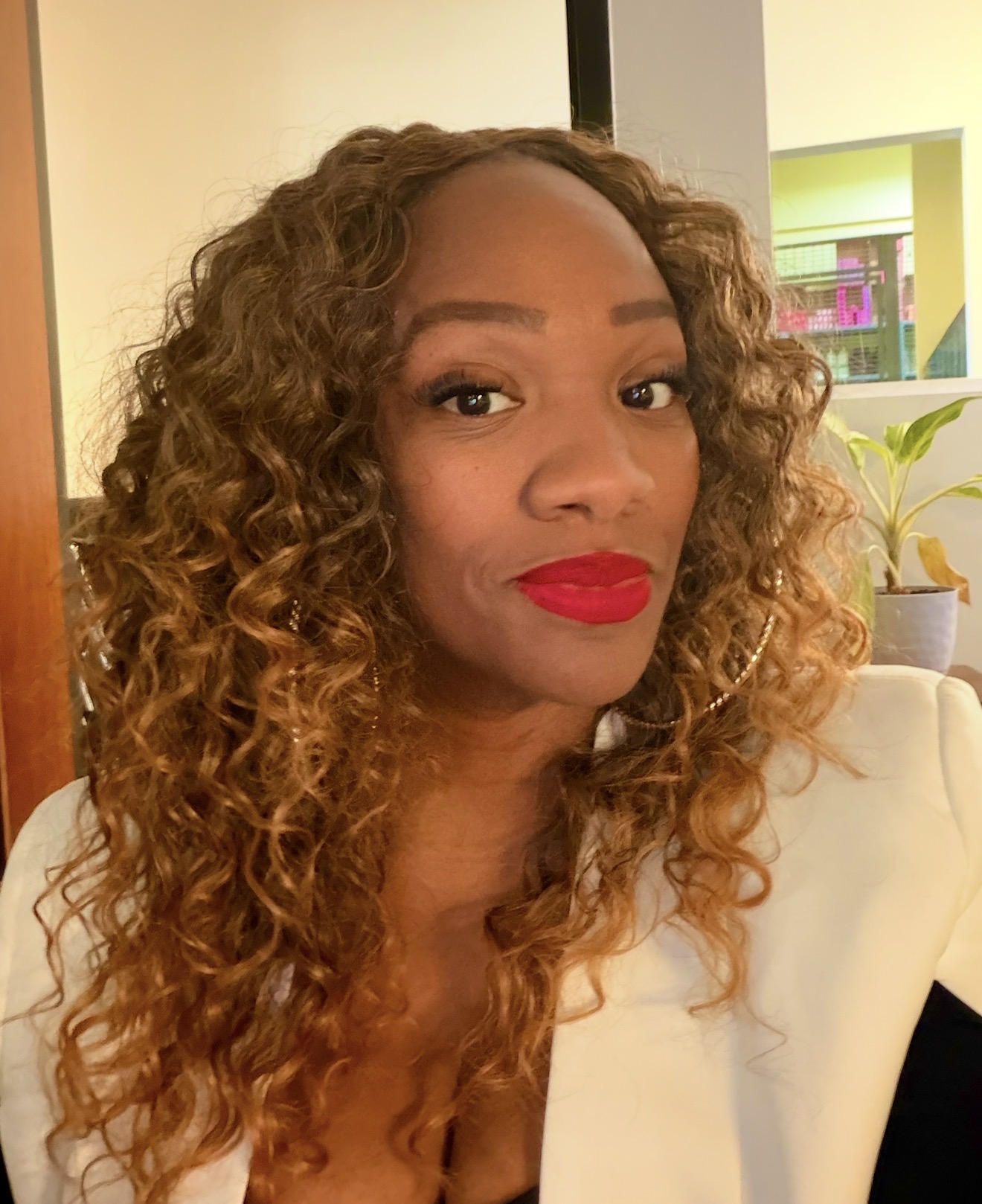 Woman with curly hair and red lipstick, wearing a white top, indoors with plants and colorful background.