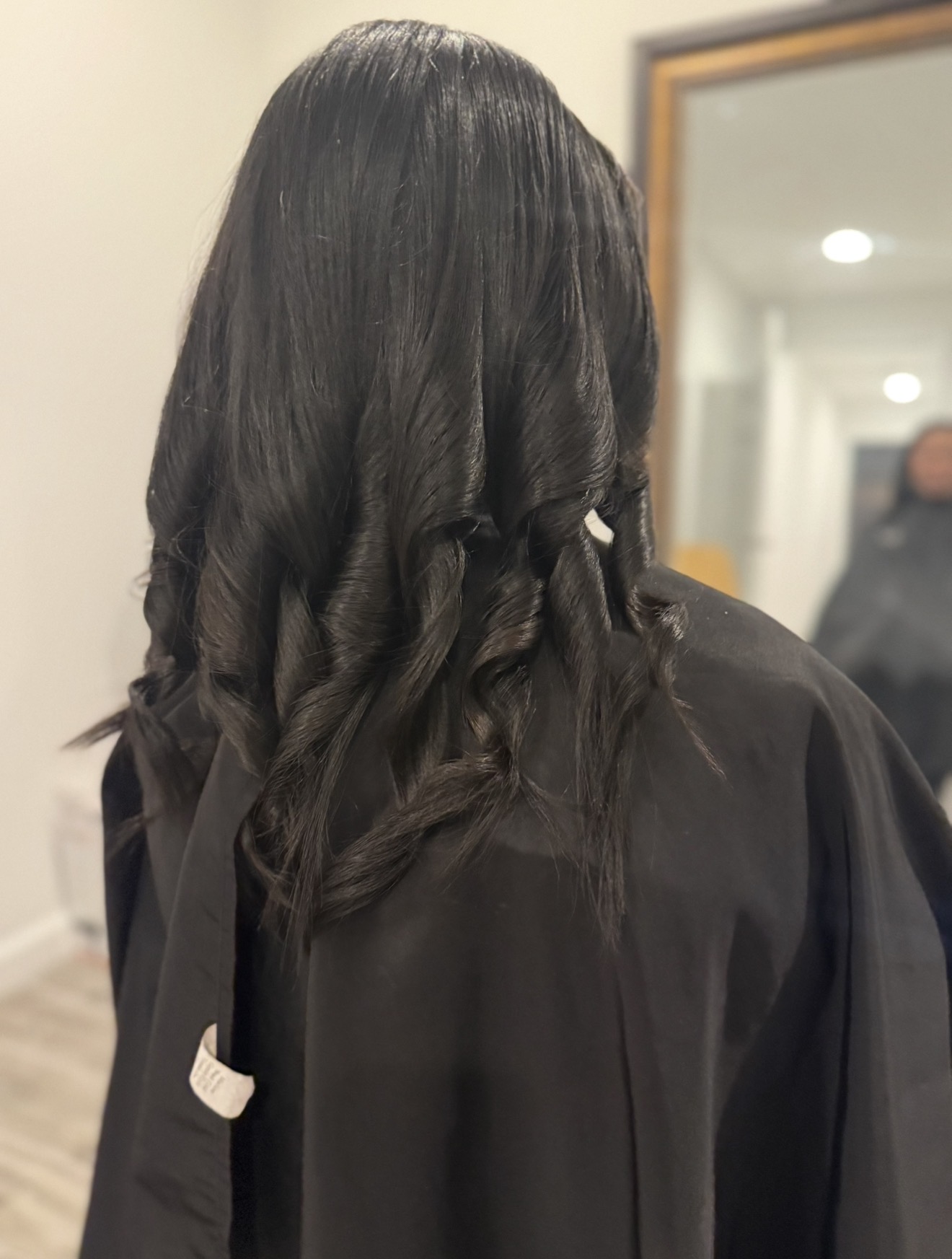 Back view of a person with long, dark, wavy hair wearing a black garment, in an indoor setting.