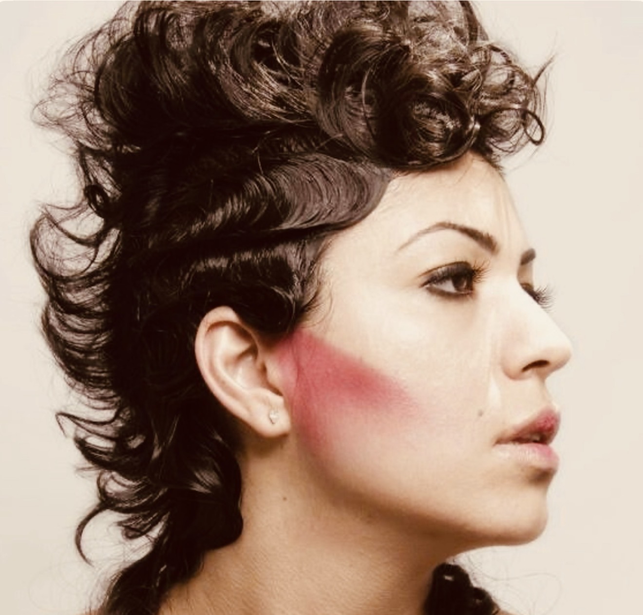 Side profile of a woman with short, curly hair and makeup, against a plain background.