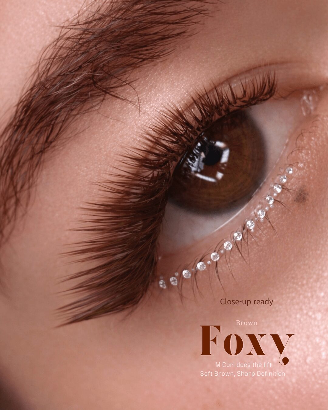 Close-up of a brown eye with long eyelashes and decorative rhinestones below, with text 'Foxy' and makeup details.