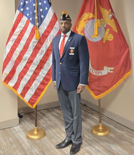Man in formal uniform standing between American and Marine Corps flags indoors, facing forward.