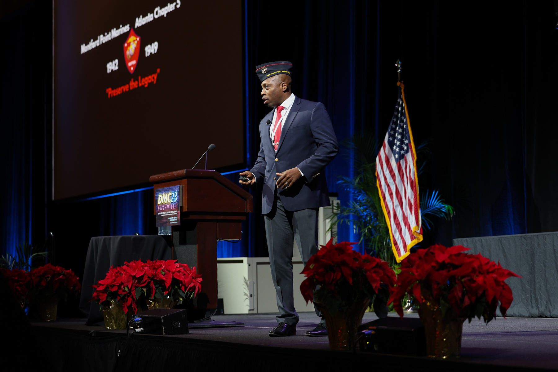 Man in suit and tie speaking at podium on stage with American flag, red flowers, and a large screen behind him.