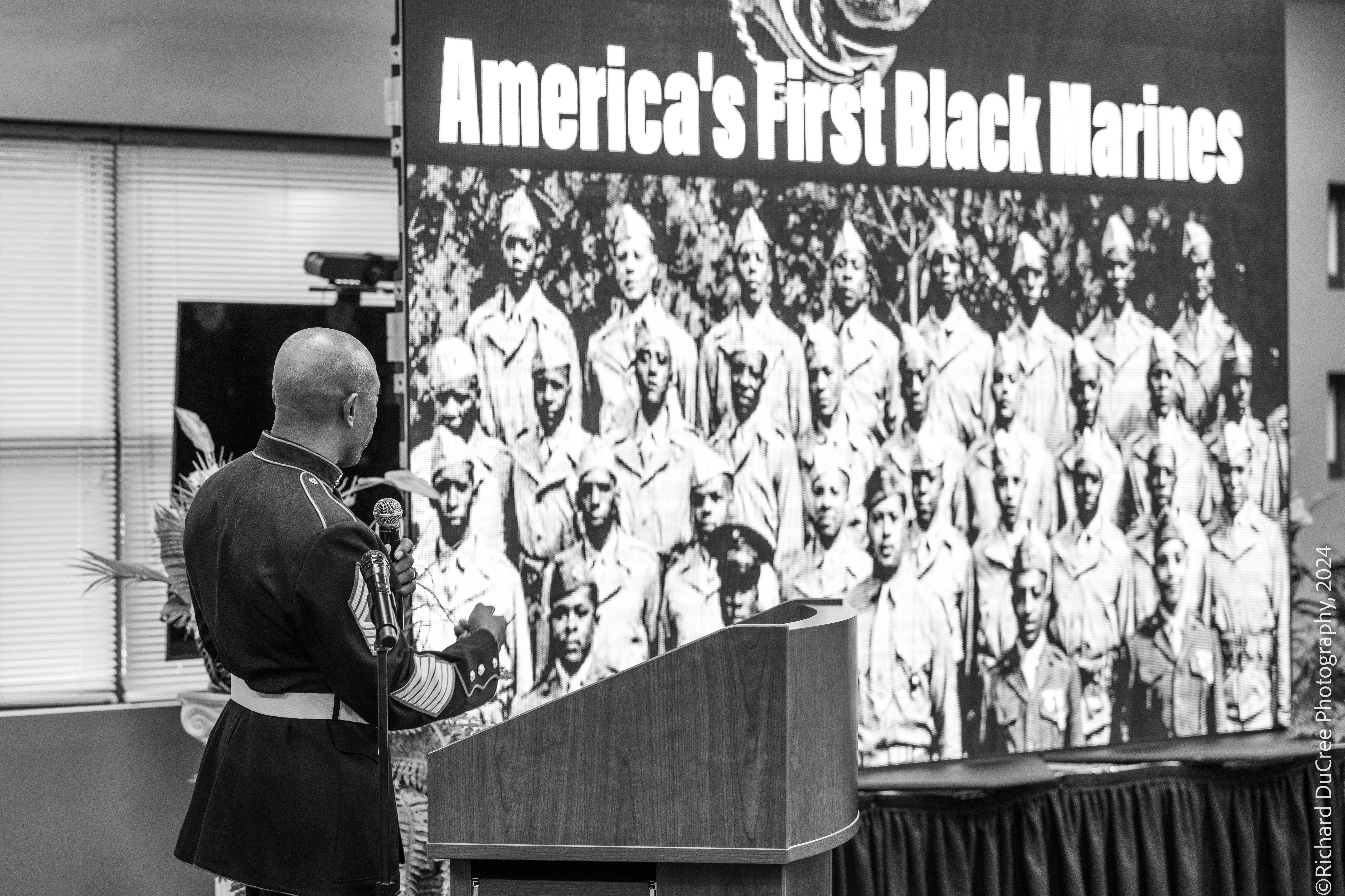 Bald man in military uniform speaking in front of a large screen displaying a group photo of Black Marines with the text 'America's First Black Marines'.