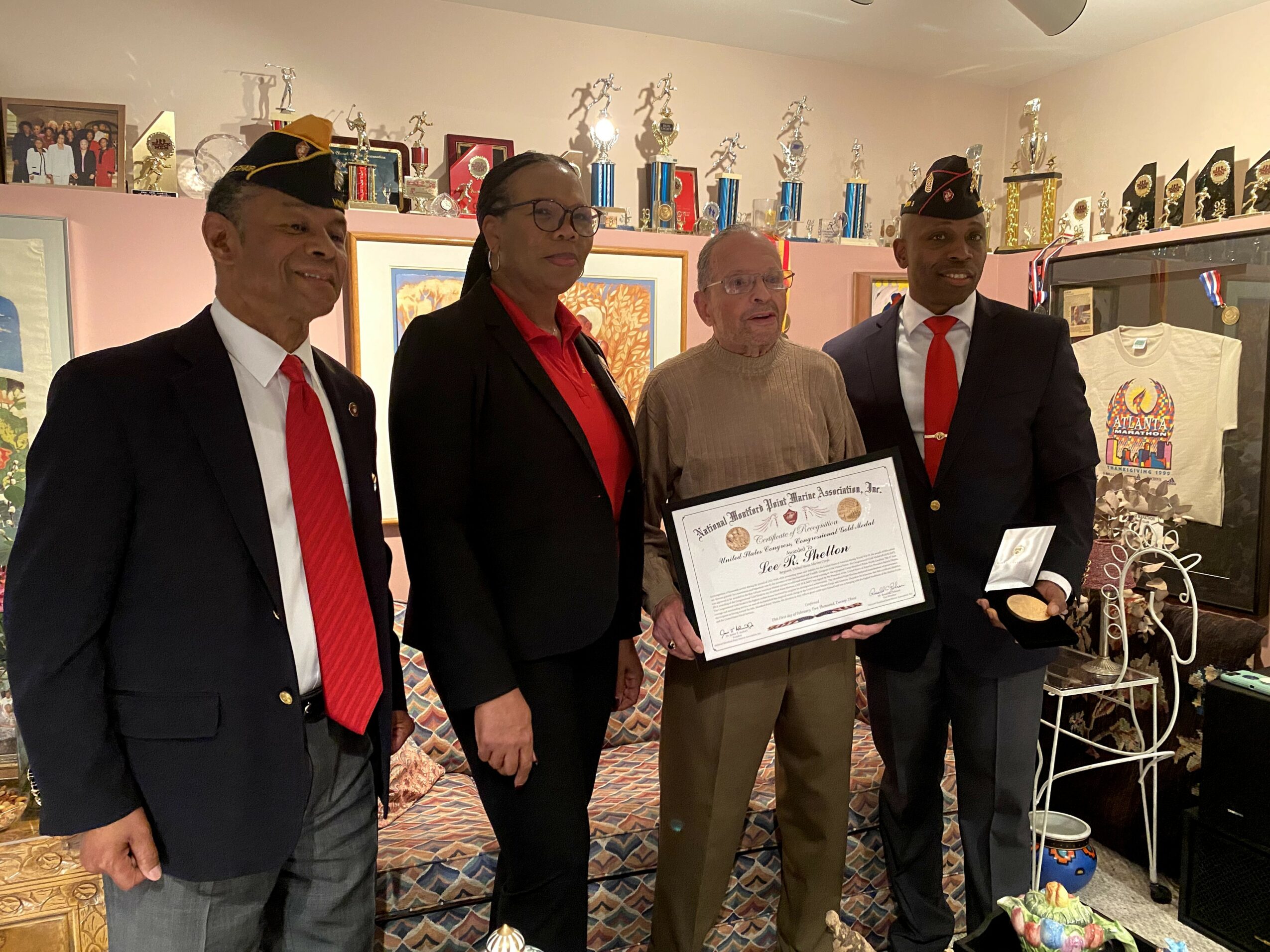 Four people standing indoors, one holding a framed certificate, trophies on shelf behind, casual and formal attire, smiling.