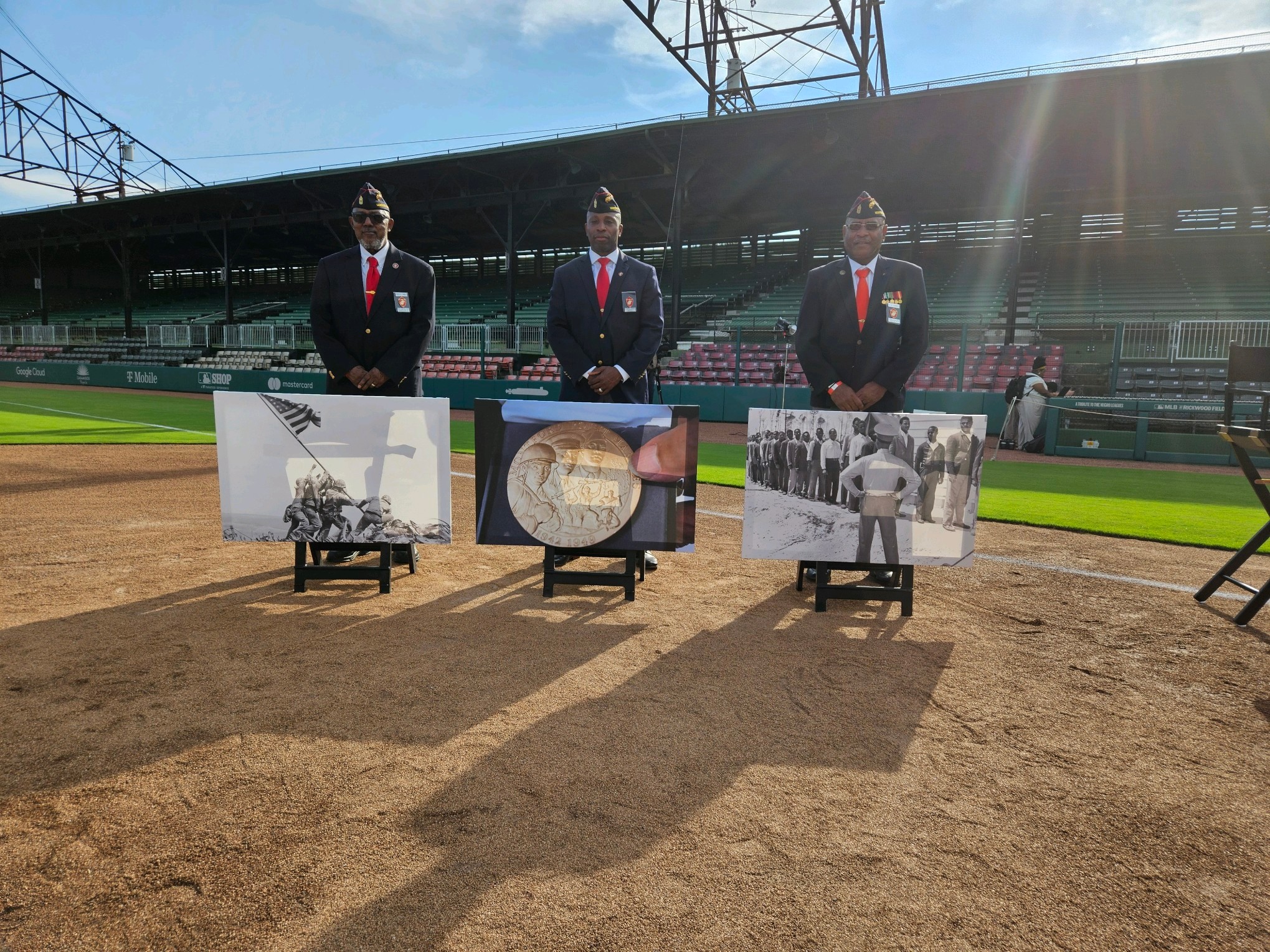 Three people in suits stand behind three framed photographs on a baseball field, with stadium seating and a blue sky in background.