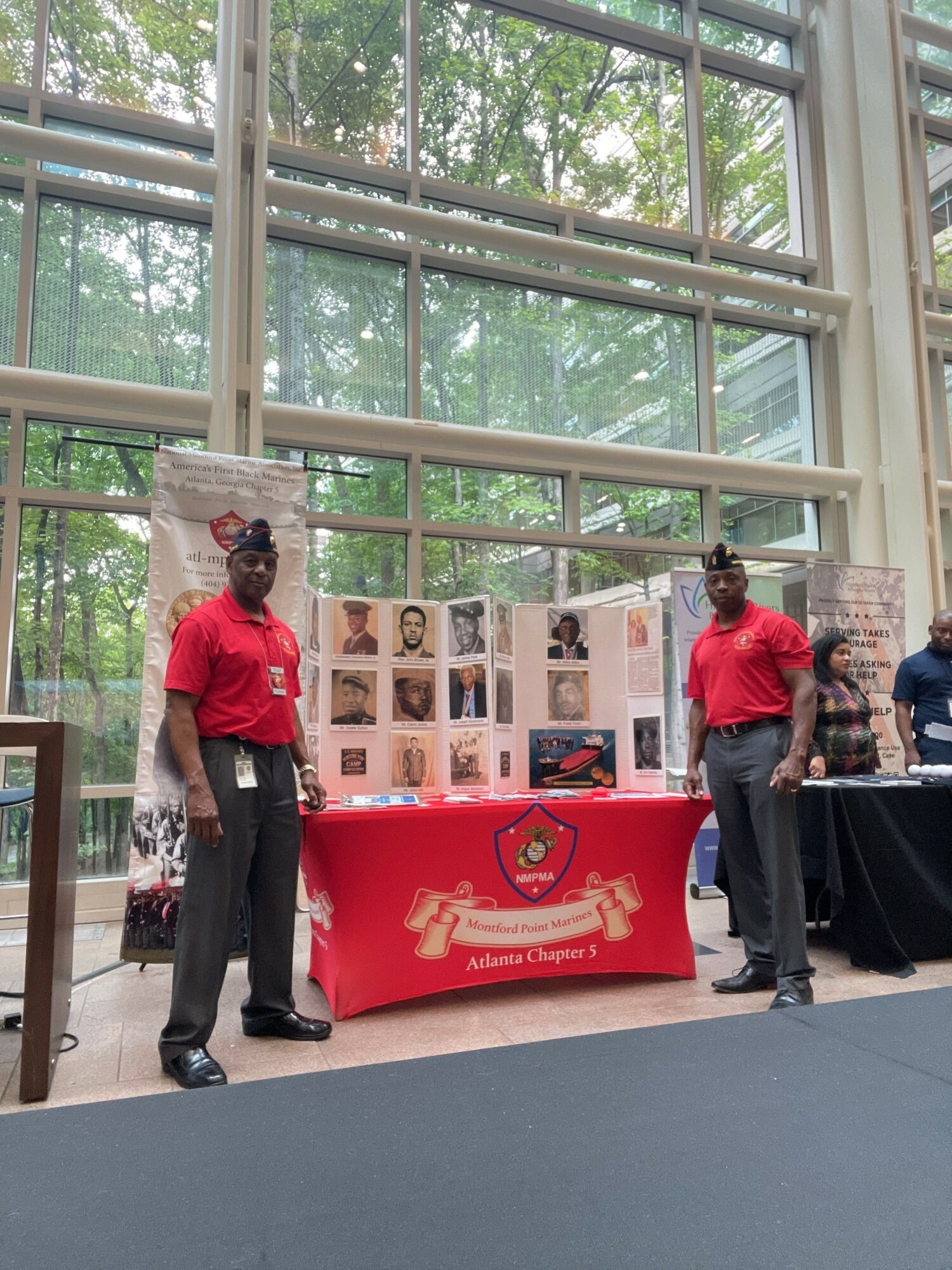 Two people in red shirts stand near a display booth with photos and a red tablecloth, inside a glass-walled building.