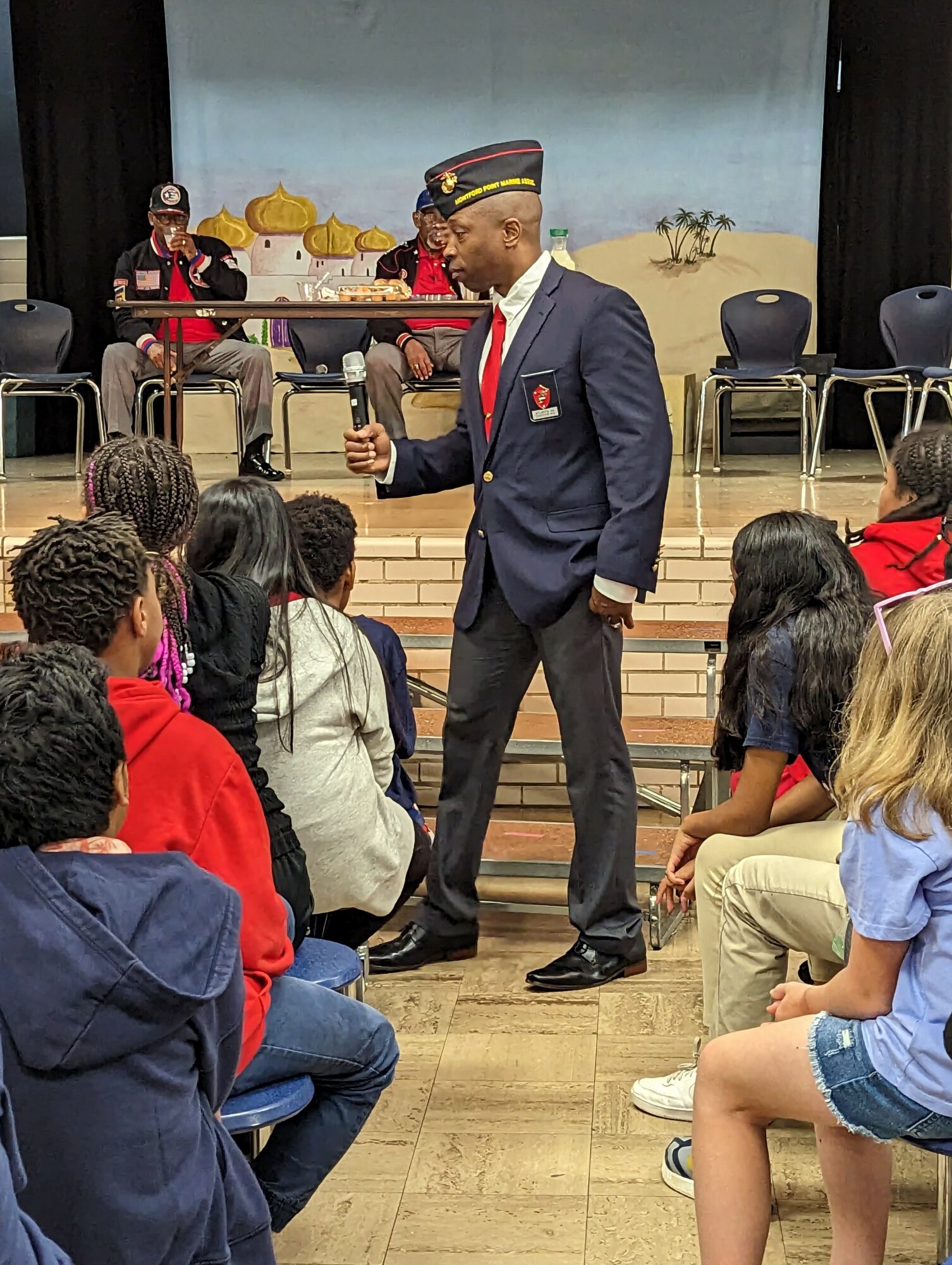 Man in uniform speaking to children seated on steps in a room with a stage backdrop of a beach scene.
