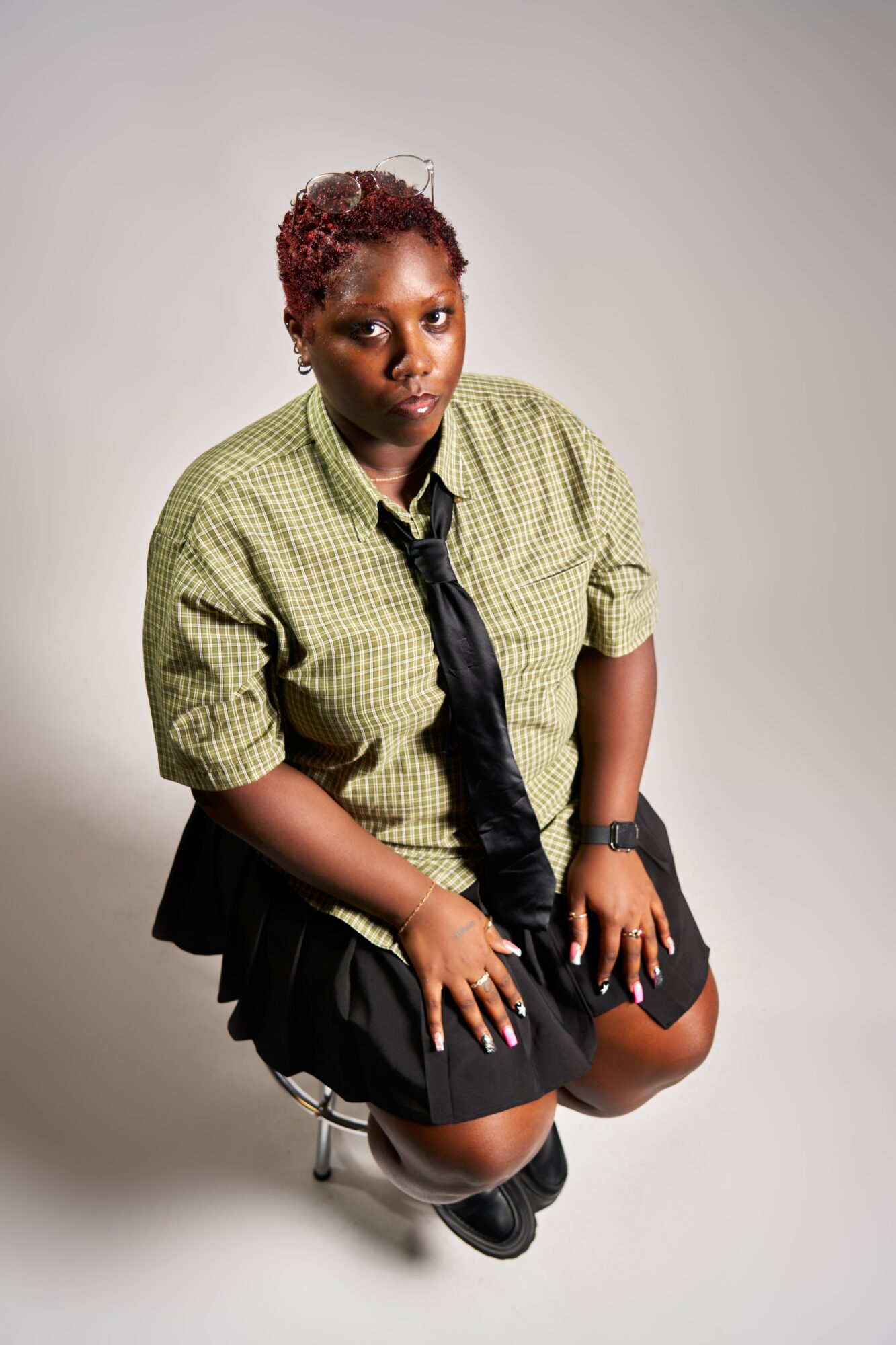 Person with short curly hair wearing a green checkered shirt, black tie, and black skirt, sitting on a stool against a plain background.