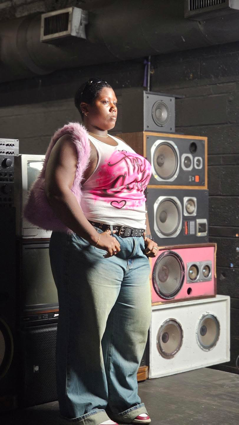Woman standing in front of stacked speakers, wearing a pink and white top, jeans, and a pink fuzzy jacket, in a dark room.