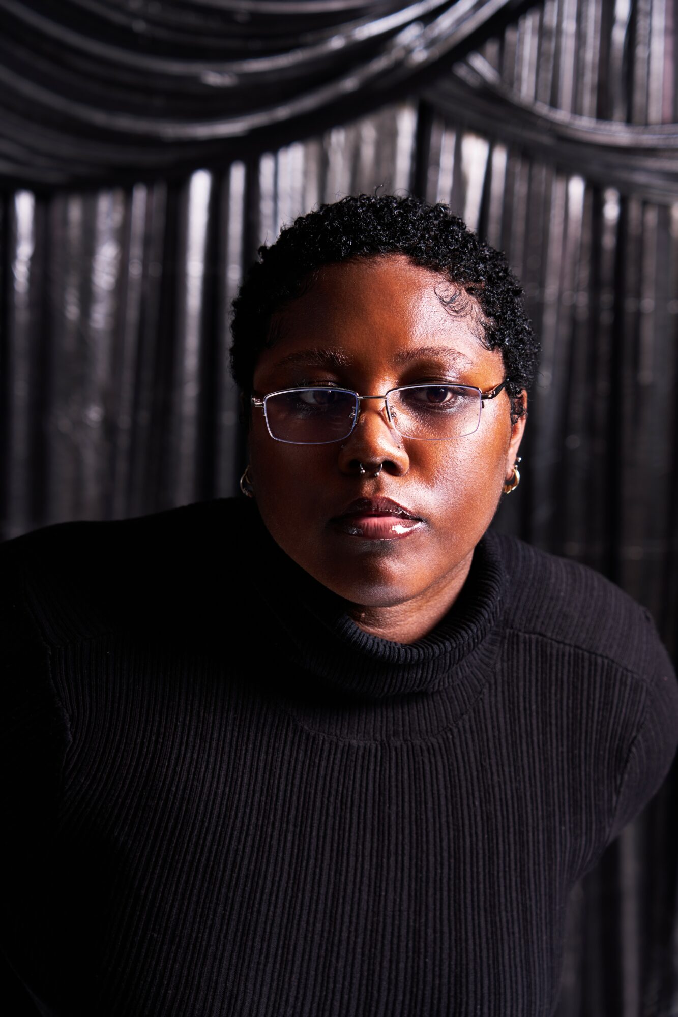 Person with short curly hair and glasses, wearing a black top, against a dark curtain background.