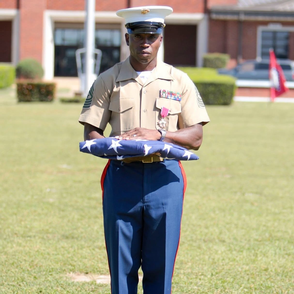 A person in military uniform holding a folded American flag outdoors, with a building and American flags in the background.