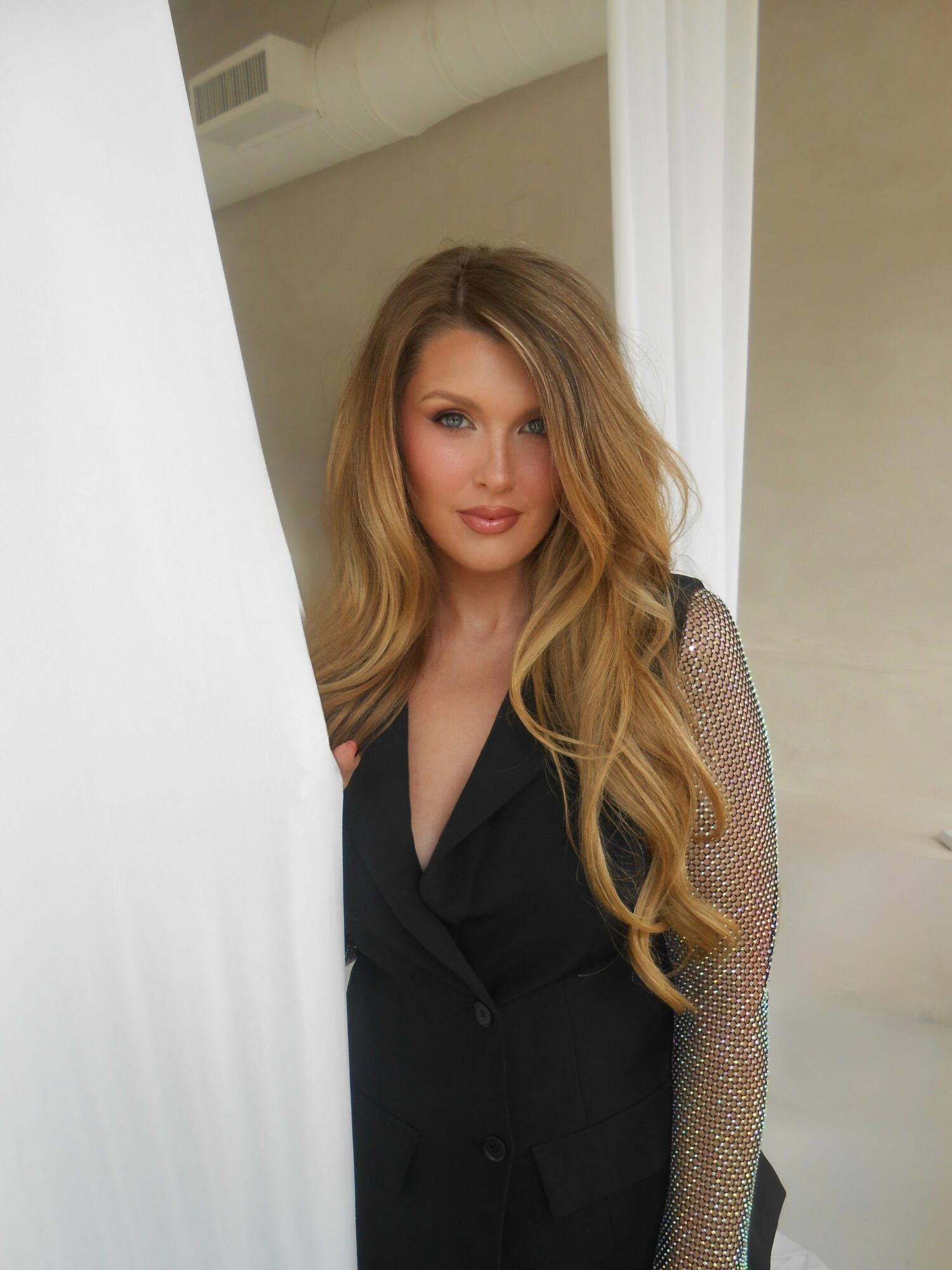 Woman with long wavy hair wearing a black blazer with sheer, sparkly sleeves, standing indoors near a white curtain.