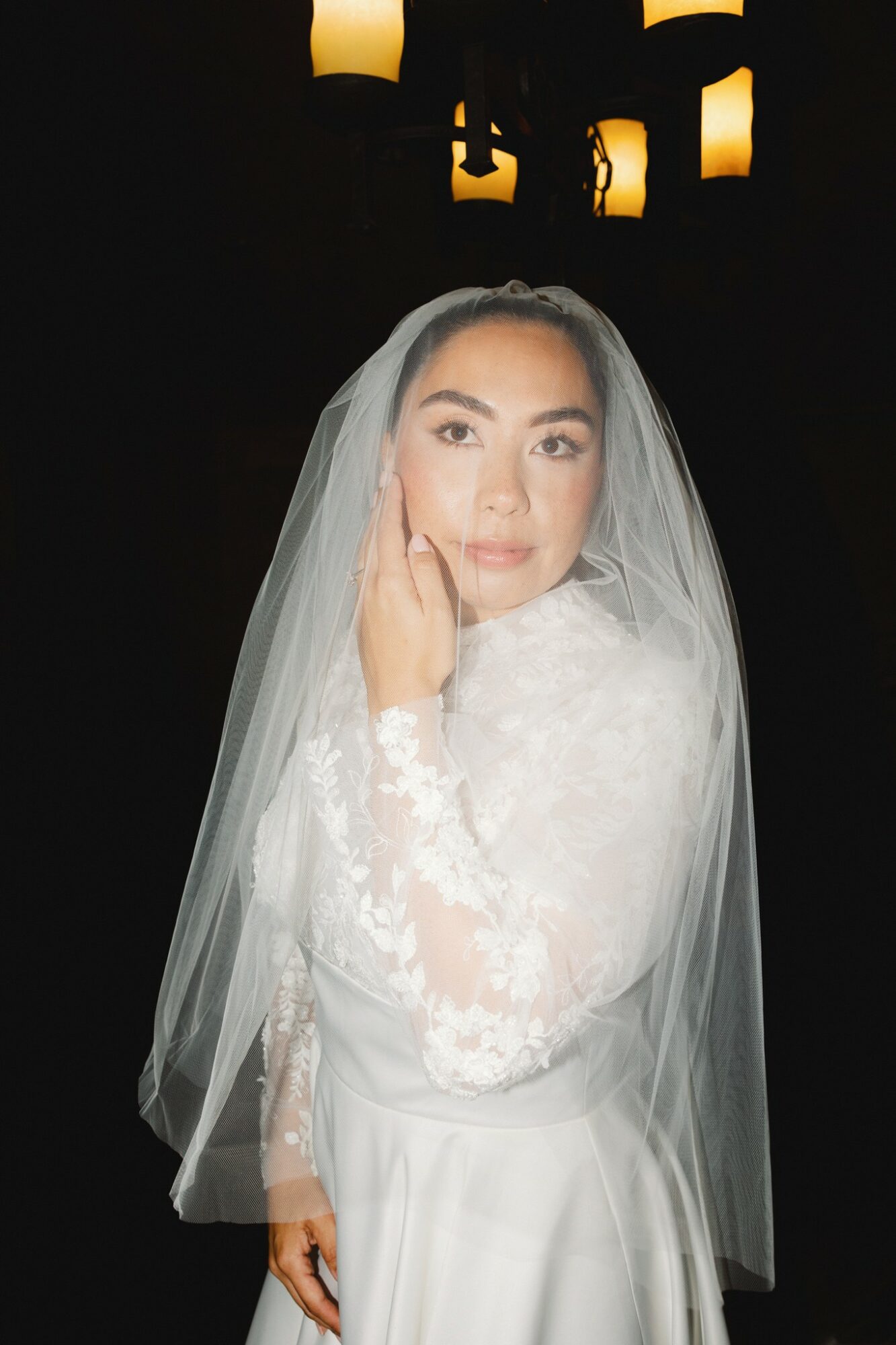 Woman in wedding dress with veil, touching face, dark background with hanging lights, soft lighting, elegant attire