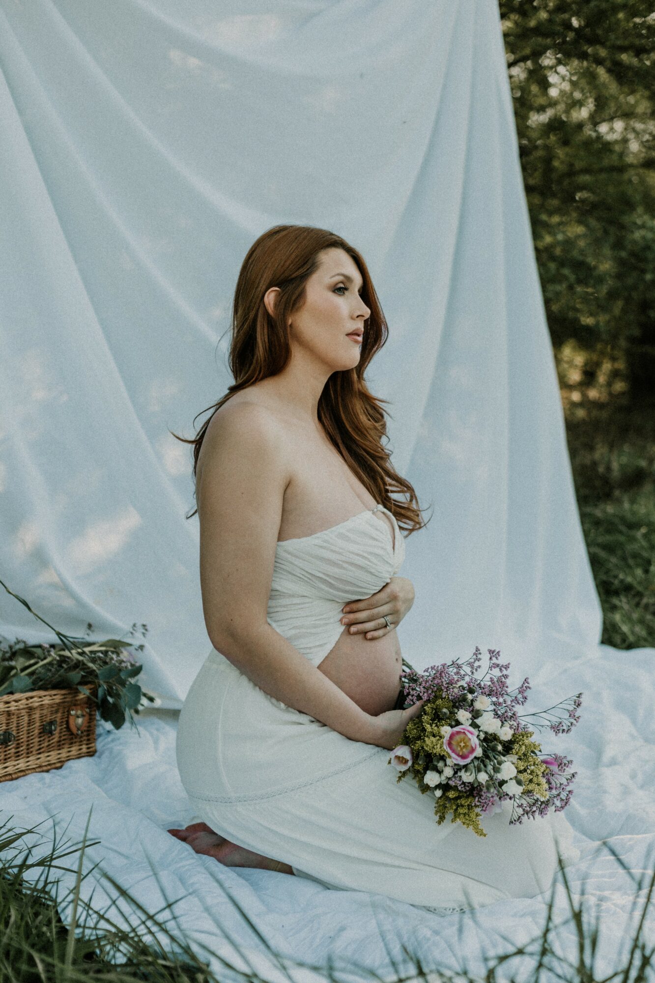 Pregnant woman with long brown hair in a white dress sitting outdoors with a bouquet of flowers, against a light fabric backdrop.