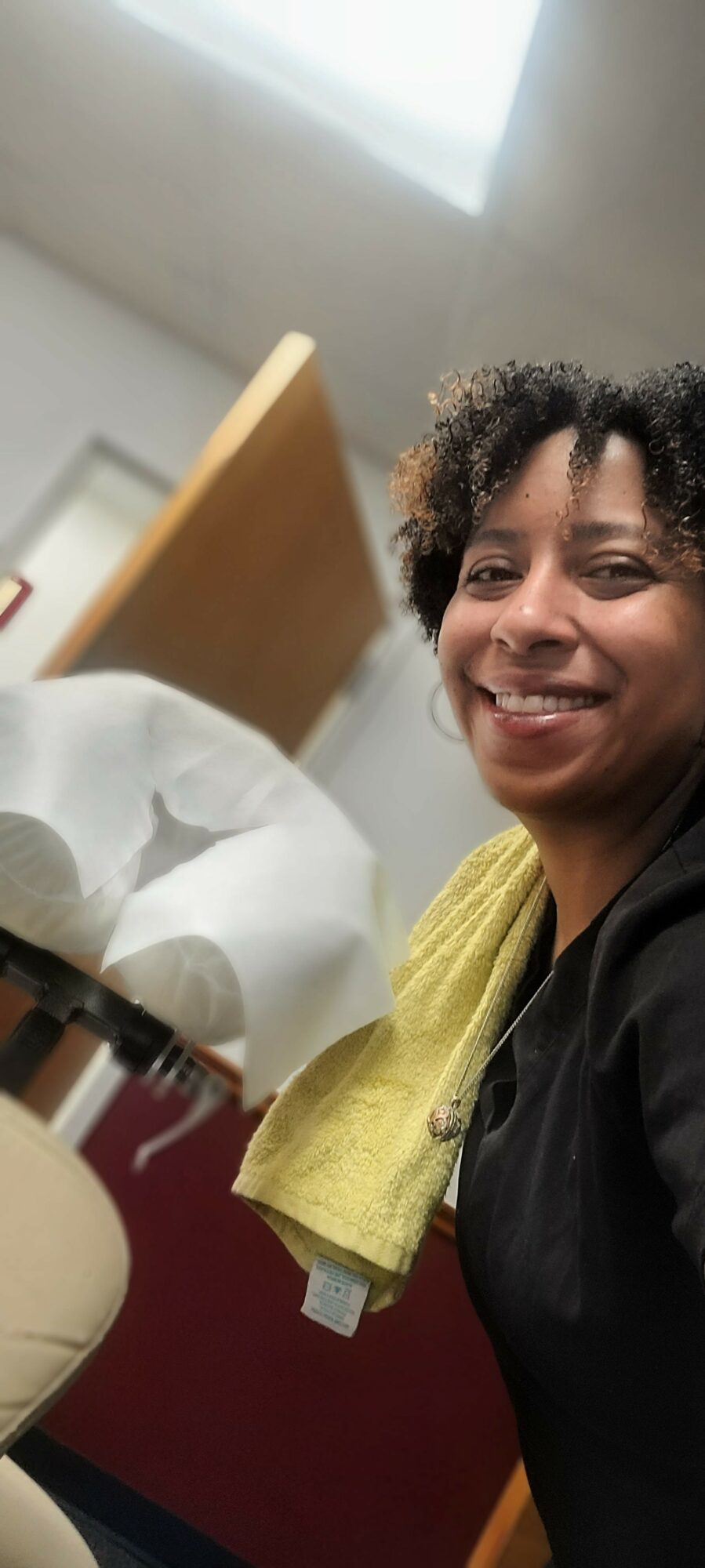 Smiling woman with curly hair in a room, with a towel over her shoulder and a bed in the background.