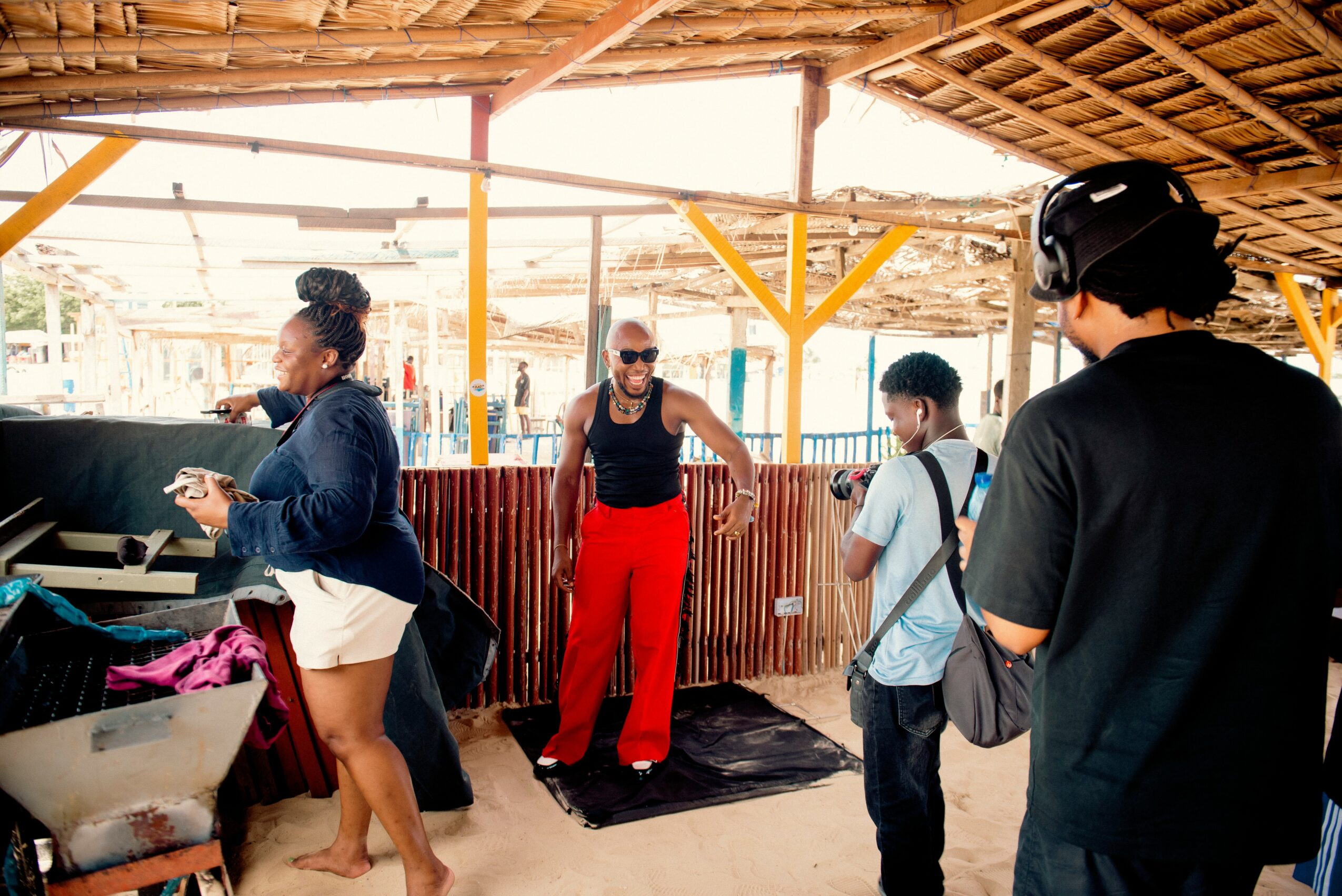 Four people under a wooden roof, one person playing piano, others standing and talking, with a yellow structure in the background.