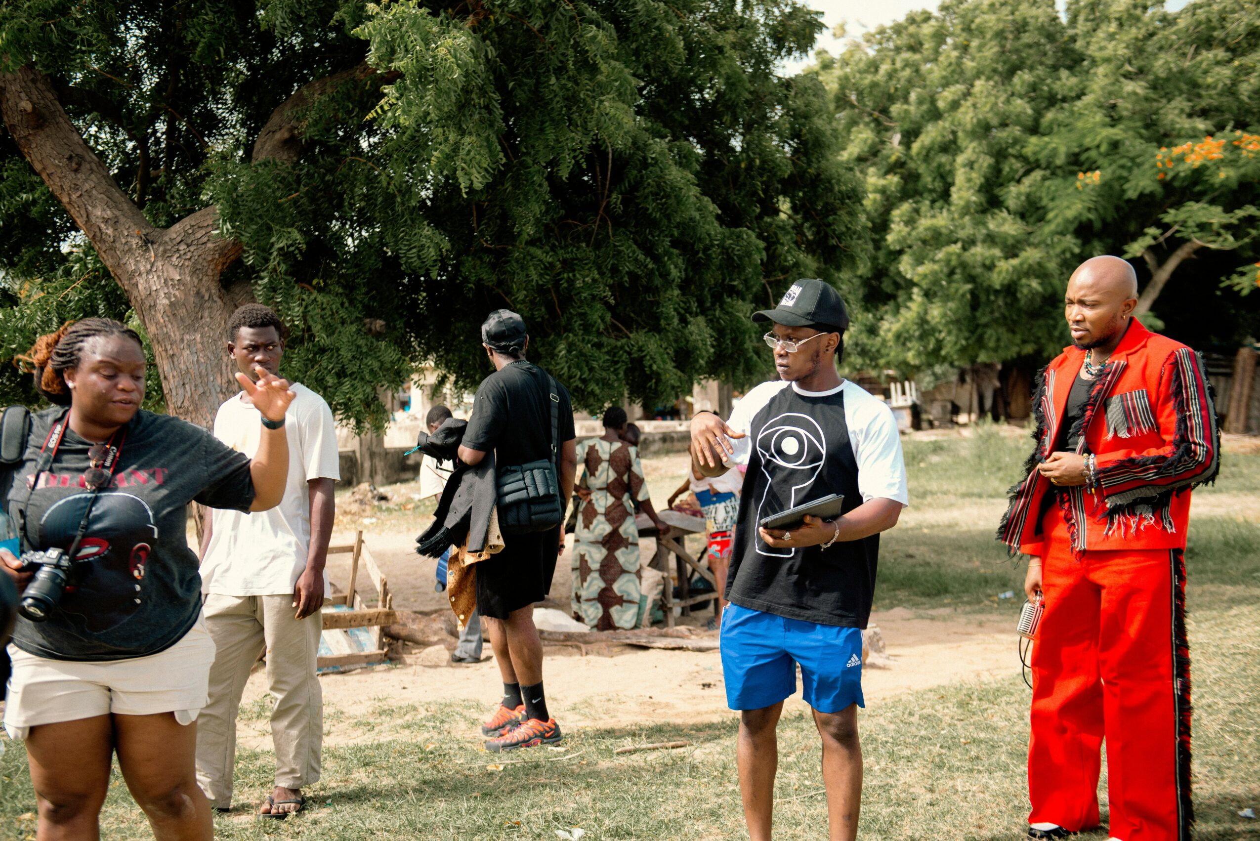 Group of people standing outdoors near trees, some talking and others looking around, in a park or open area.