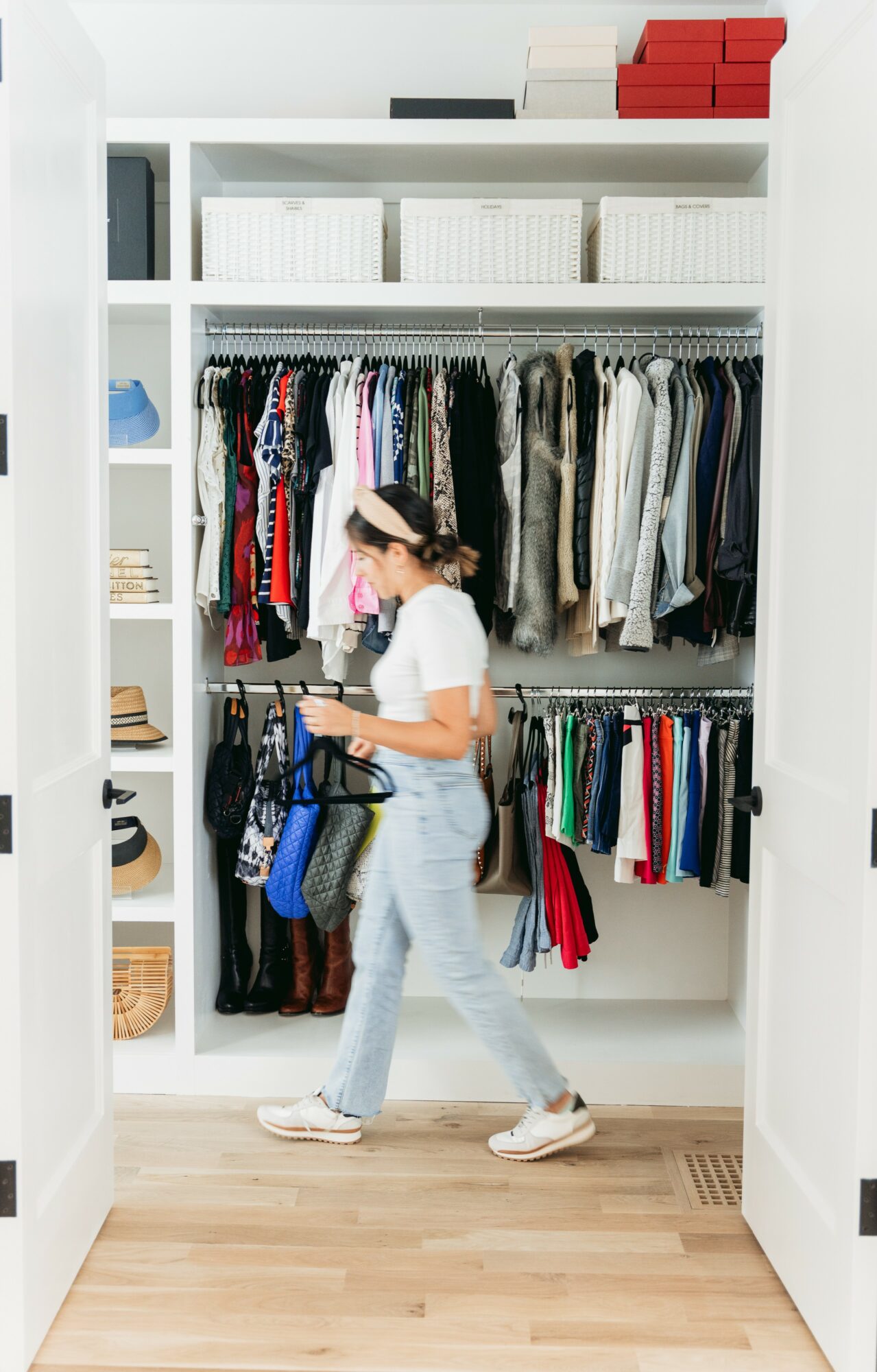 Person browsing clothes in a white closet with shelves and hanging rods, wearing a white shirt, light jeans, and a pink headband.