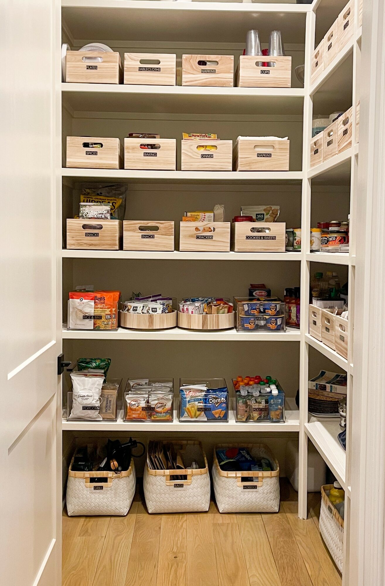 Closet with shelves filled with labeled storage boxes and baskets, containing various household items, on a wooden floor.