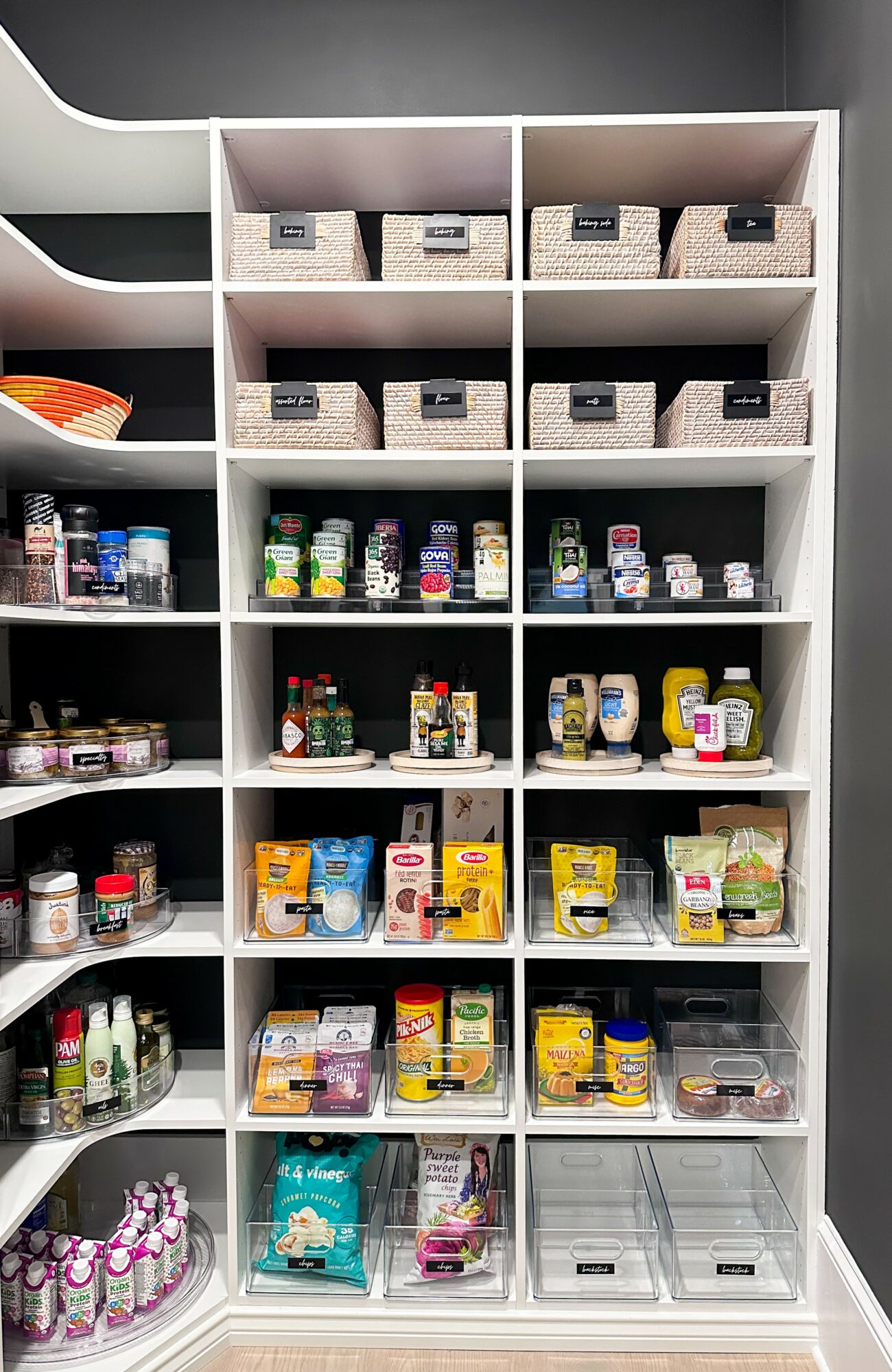 White shelving unit with storage baskets on top and various food and condiment containers on middle and lower shelves.