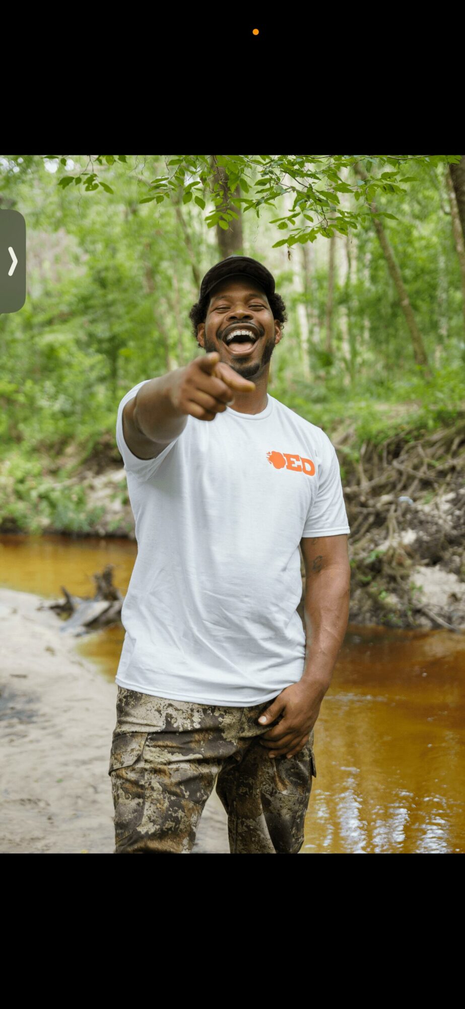 Man smiling and pointing outdoors near a river with green trees in background.
