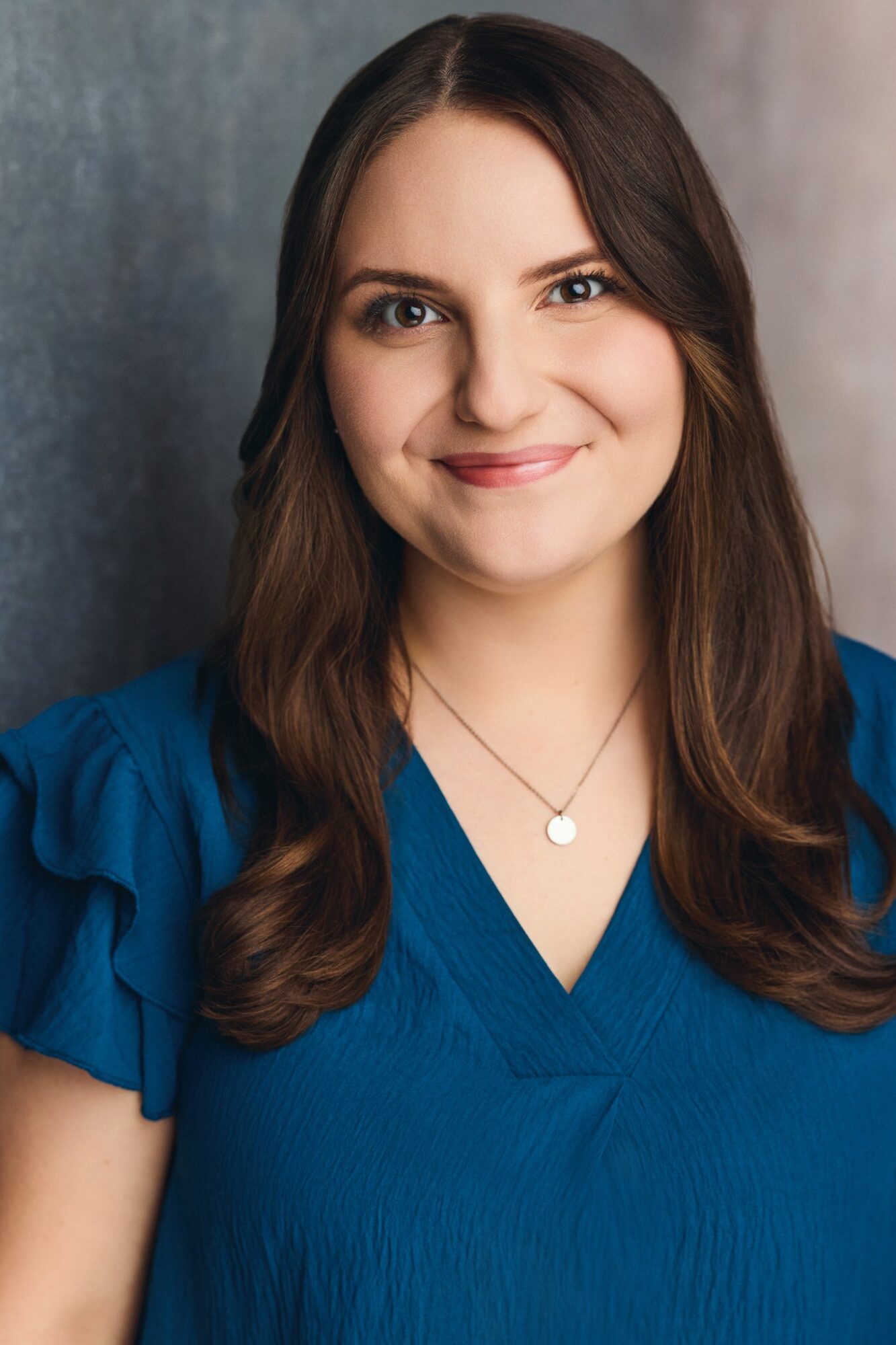 Young woman with long brown hair smiling, wearing a blue top and a necklace, against a gray background.