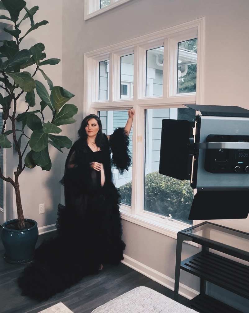 Woman with long dark hair in black dress standing near large window, with plant and equipment in room.