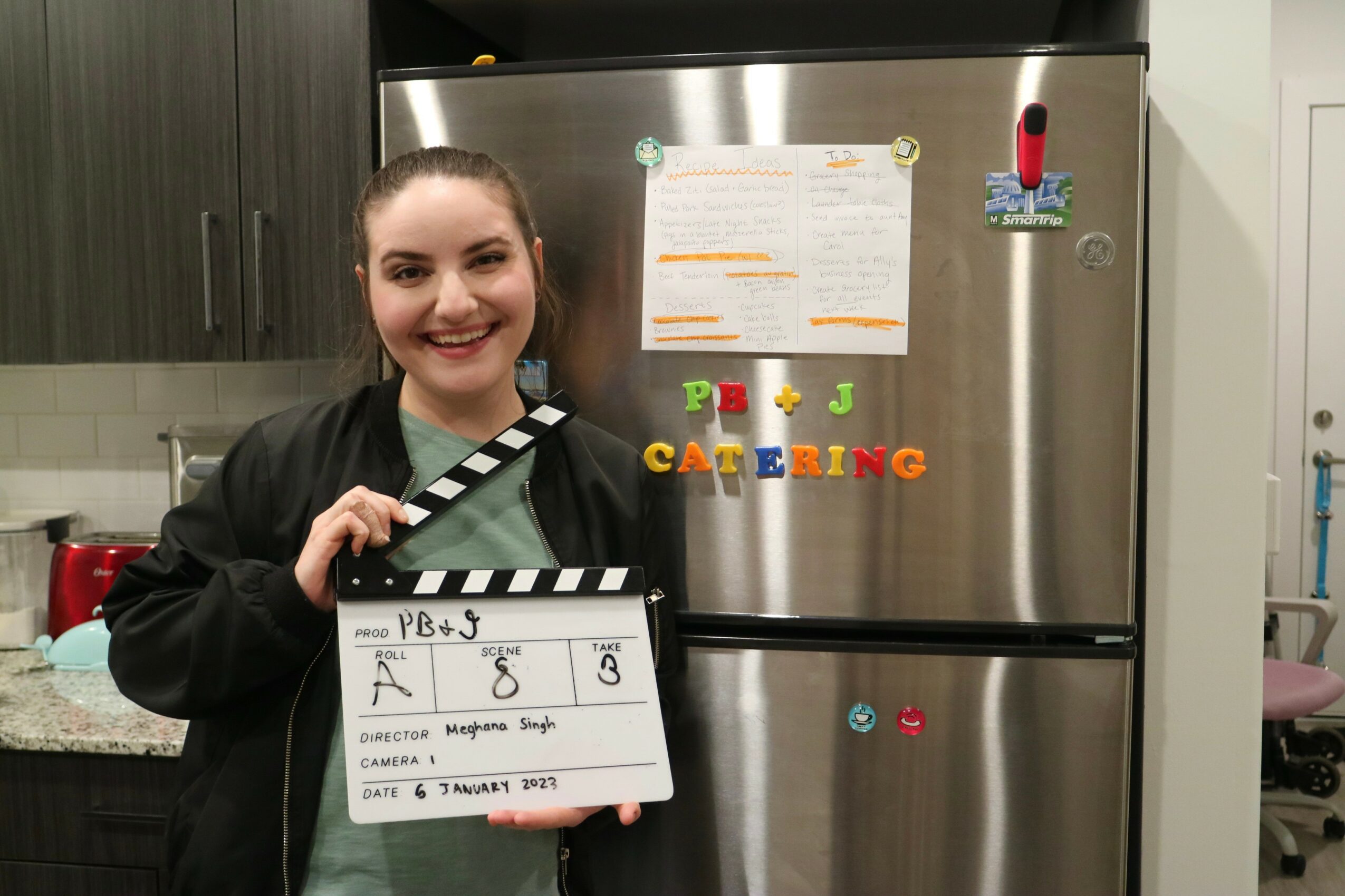 Young woman holding a clapperboard in a kitchen, smiling, with a stainless steel refrigerator behind her.