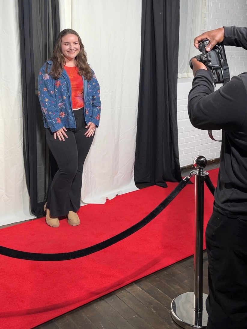Young woman with long hair smiling on a red carpet, posing for a photo, with black and white curtains behind her.