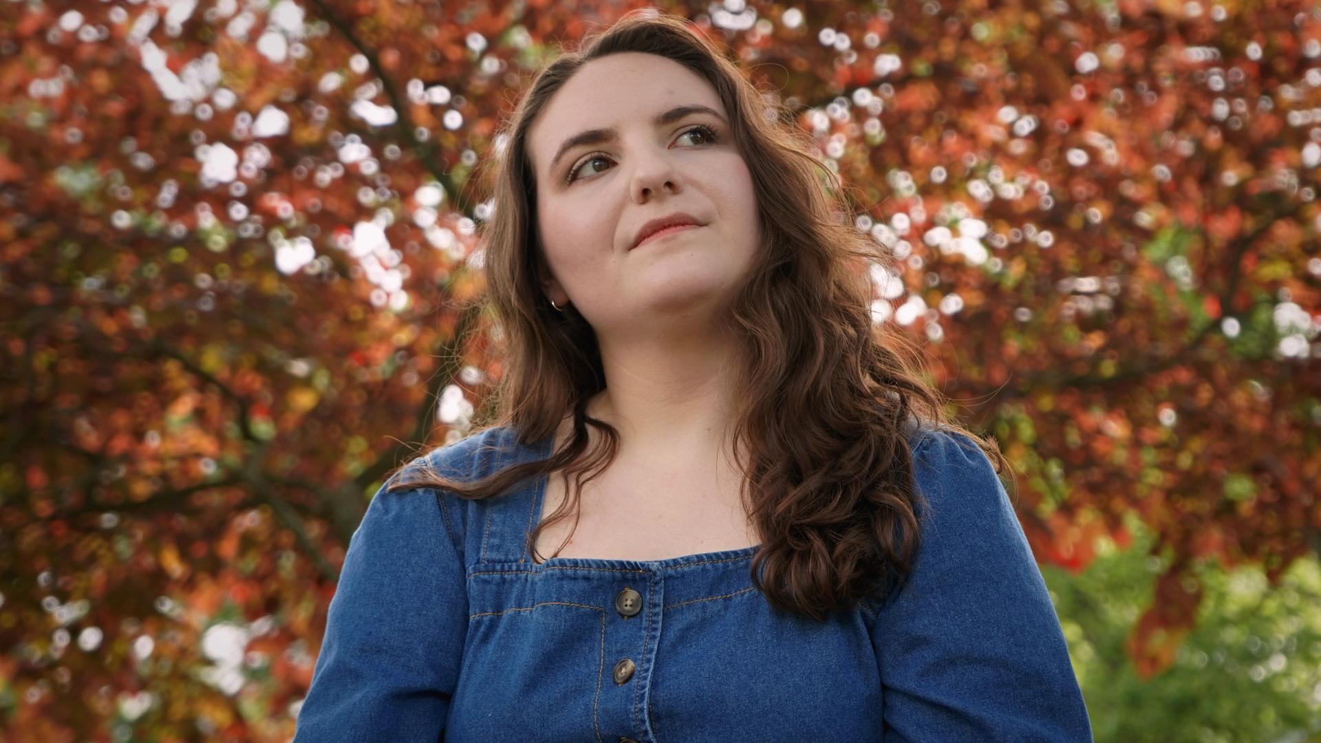 Young woman with long brown hair wearing a denim jacket outdoors with autumn trees in background.