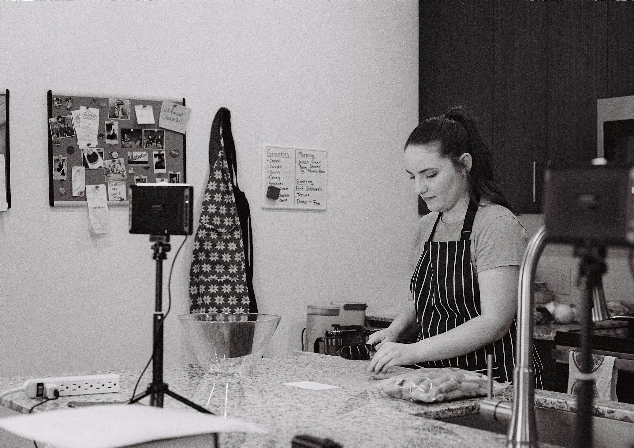 A woman in an apron prepares food in a kitchen with a bulletin board and hanging bag behind her.