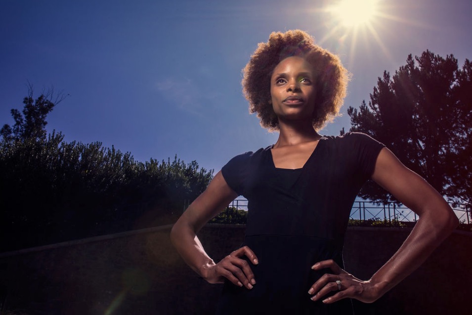 Woman with hands on hips outdoors, sun shining behind her, trees in background, clear sky.