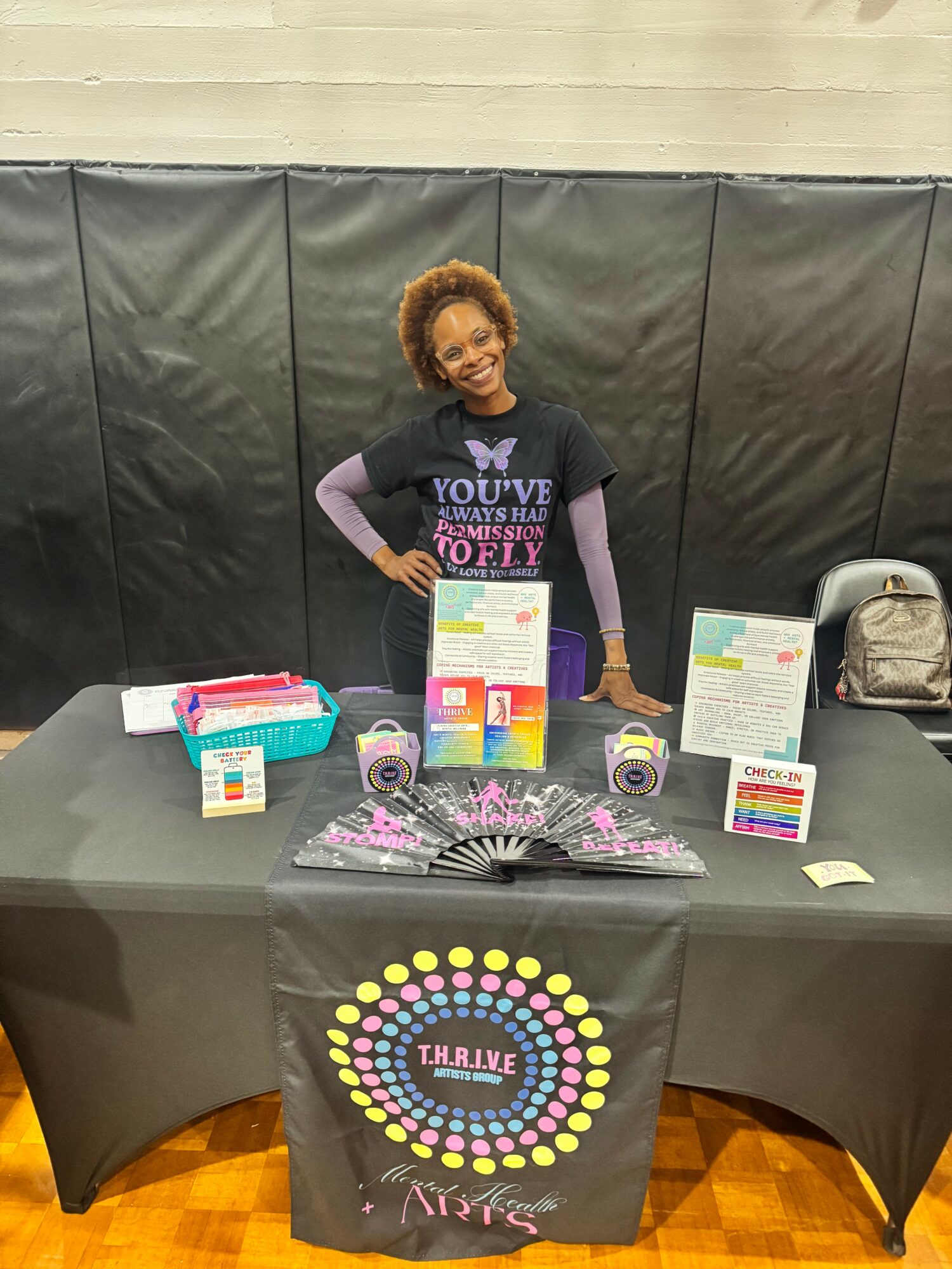 Woman standing behind a table with colorful items, black backdrop, and a logo on the tablecloth, smiling at the camera.