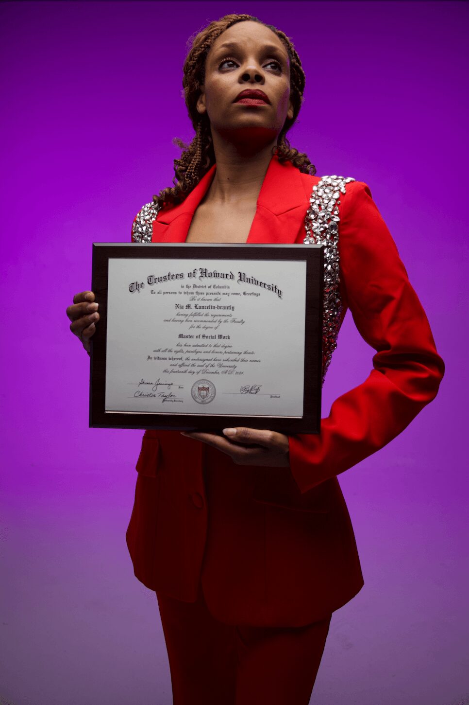 Woman in red suit holding a framed certificate against purple background.