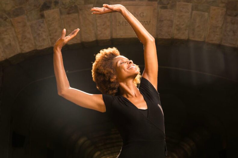 Woman with curly hair dancing with arms raised, smiling, in a dark space with stone walls behind her.