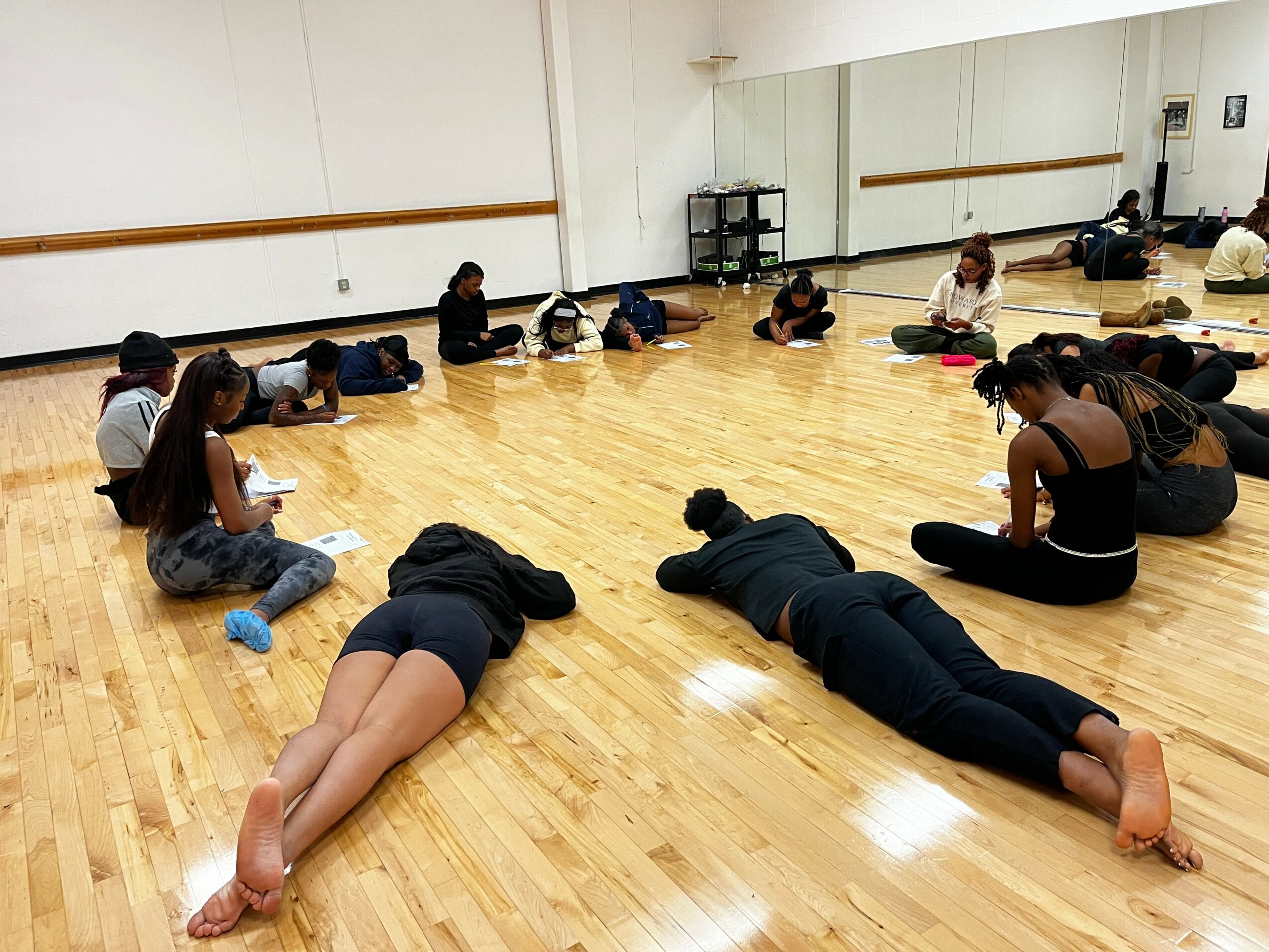 People sitting and lying on a wooden floor in a dance studio with a large mirror, some with their heads down.
