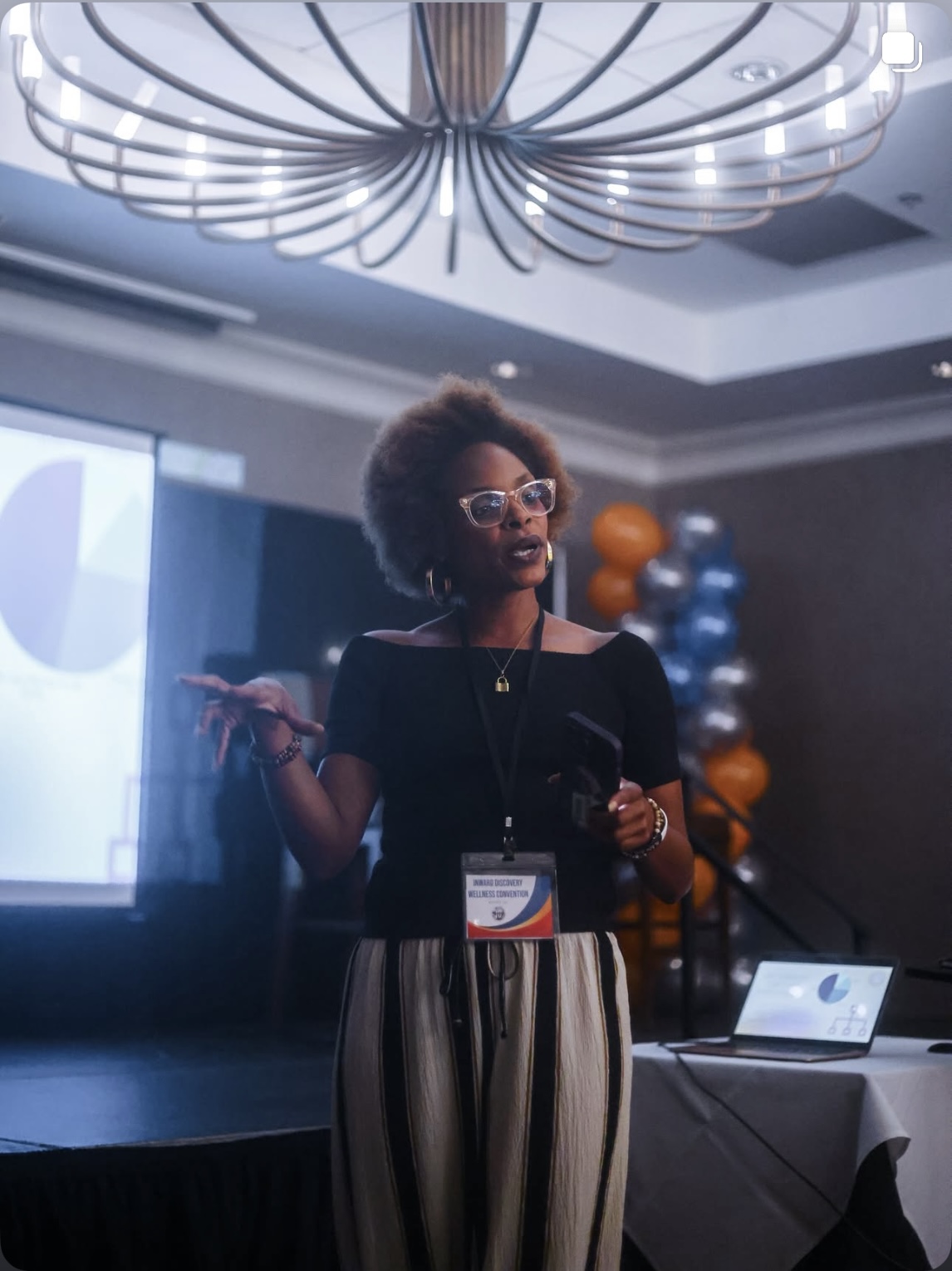 Woman with glasses and curly hair presenting at a conference, holding a remote, with a screen and balloons in background.