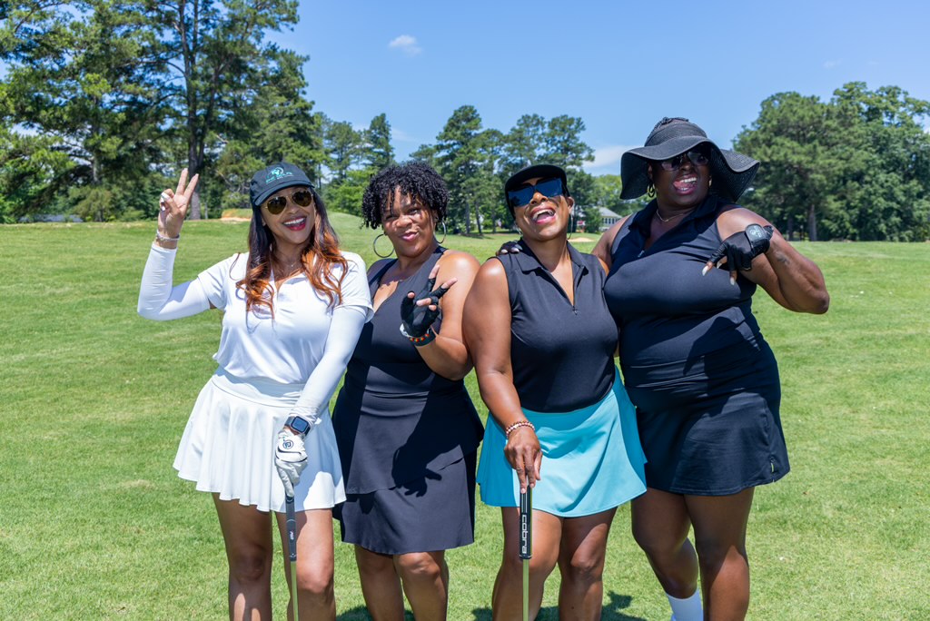 Four women standing outdoors on a sunny day, smiling, with trees and grass in the background.