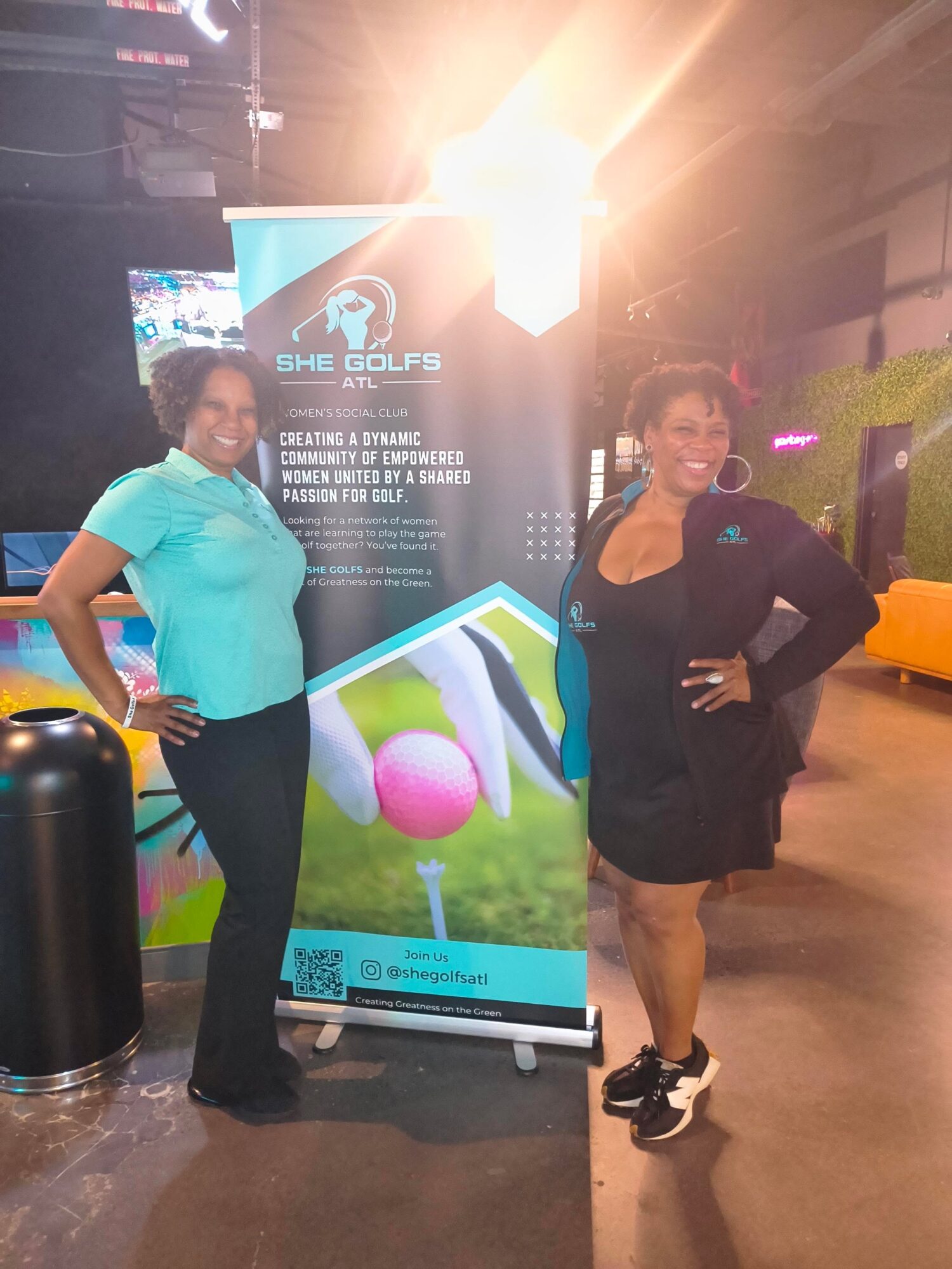 Two women standing next to a banner at an indoor event, with bright lighting overhead and a screen in the background.