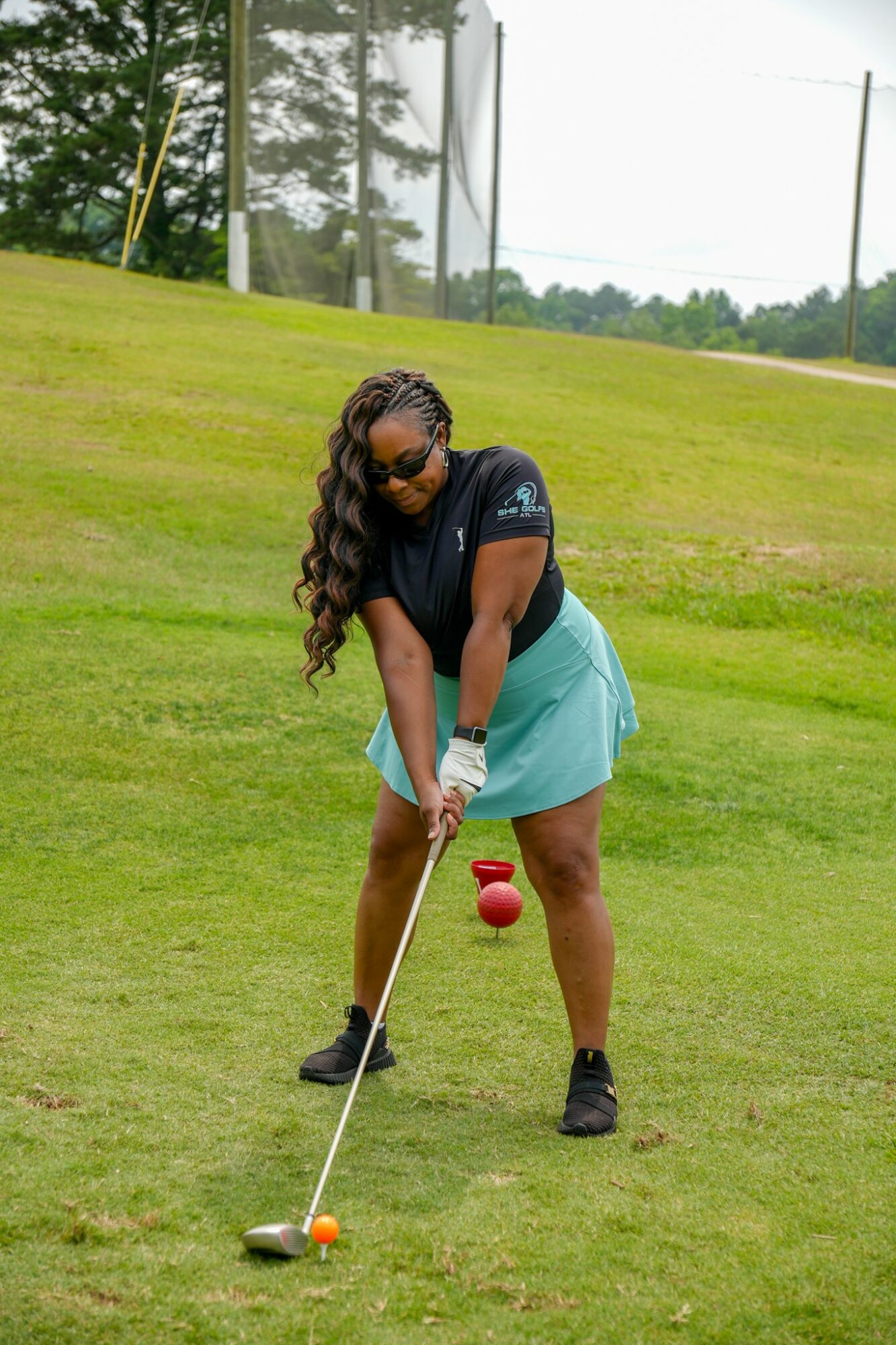 Woman preparing to hit golf ball on grassy course, wearing sunglasses, black top, and blue skirt.