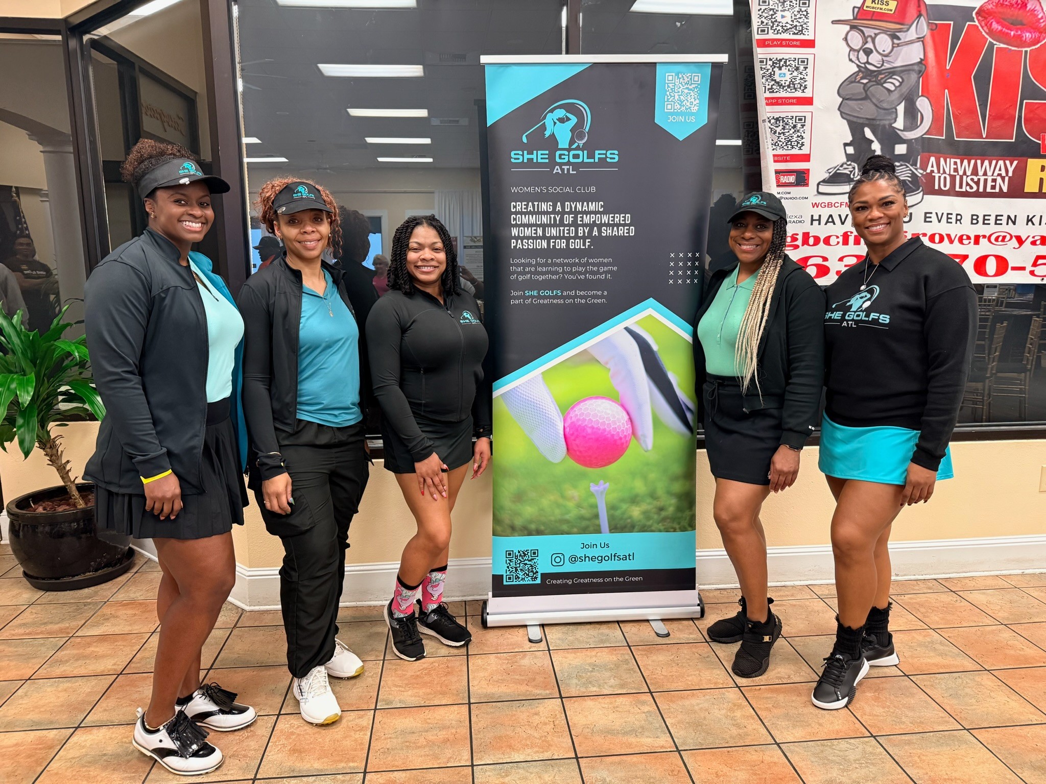 Five women stand indoors next to a vertical banner, smiling, wearing casual sportswear, with a potted plant and posters behind them.