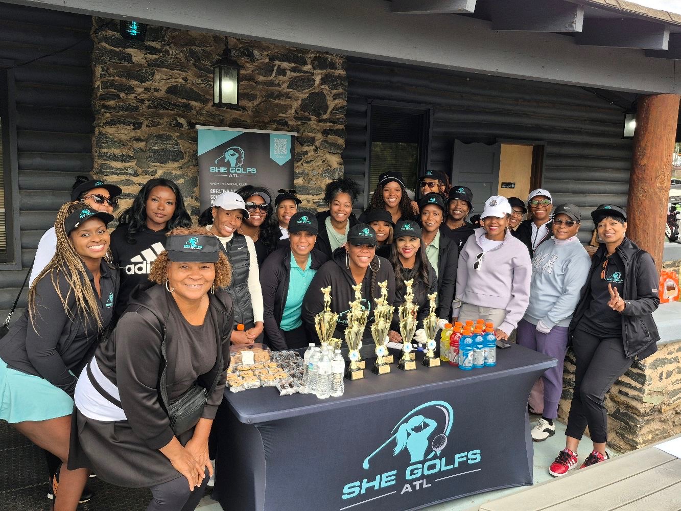 Group of women and girls gathered around a table with trophies and water bottles outside a building, smiling.