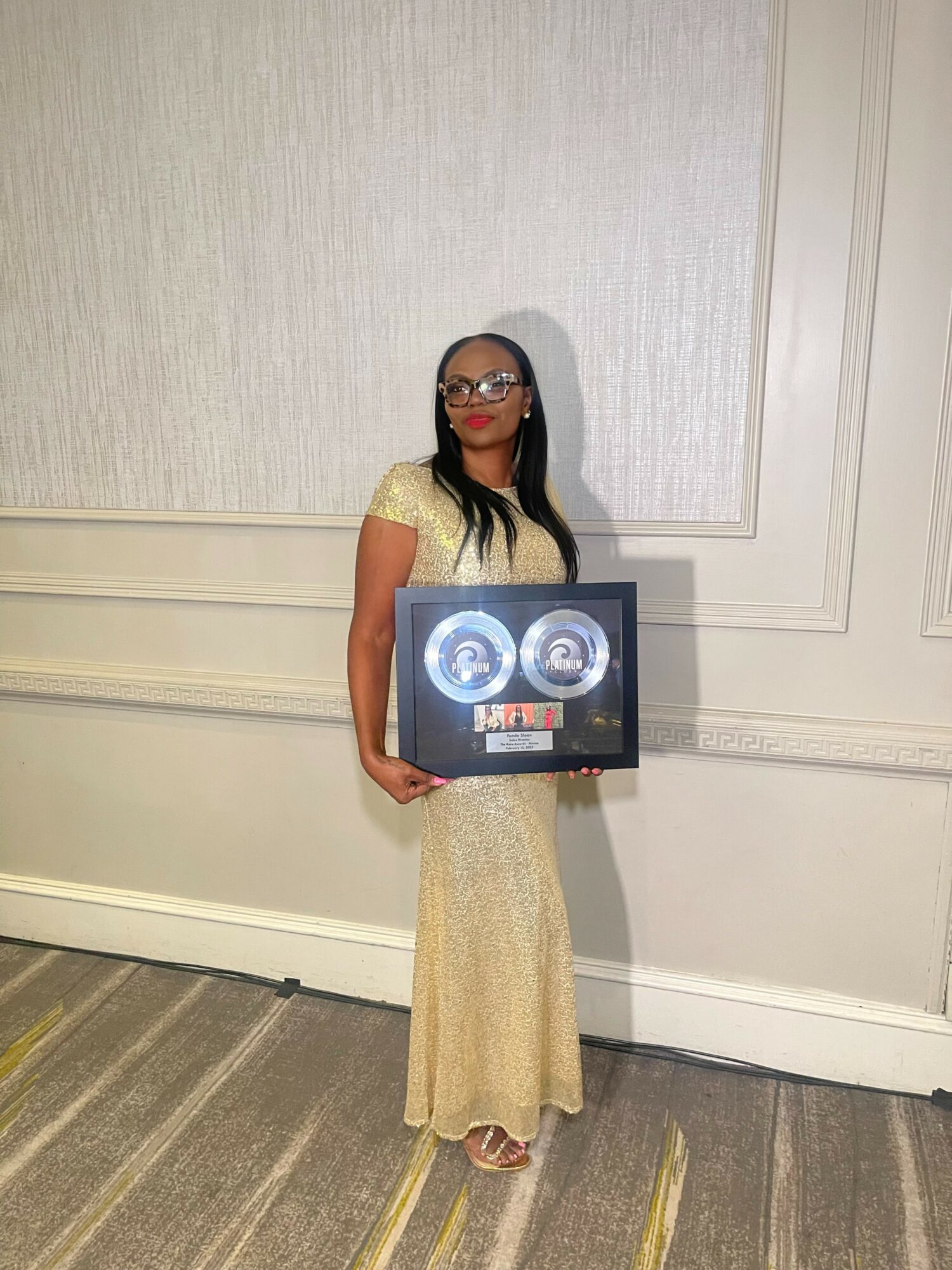 Woman in a yellow dress holding a framed award, standing against a light-colored wall.