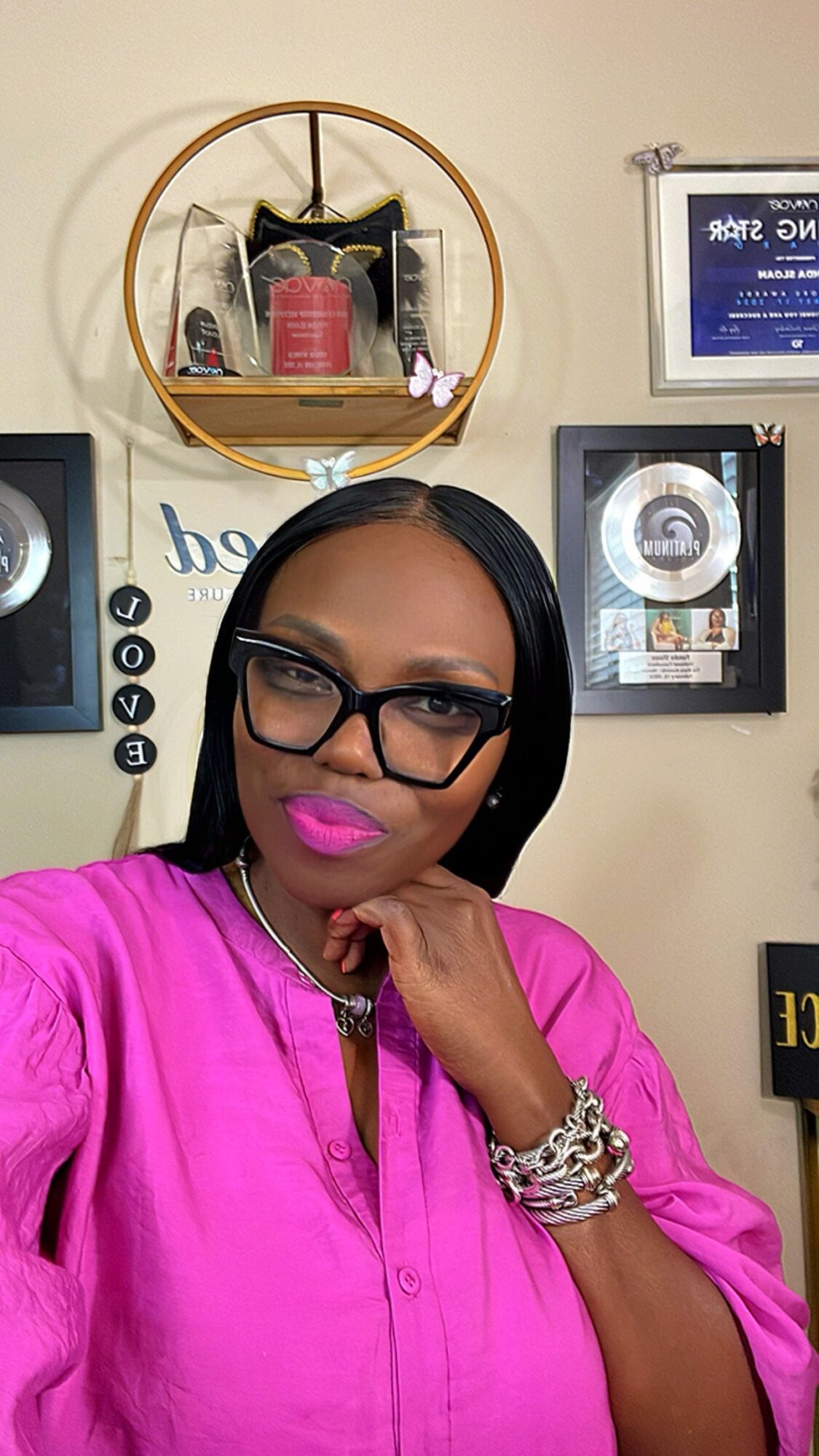 Woman with black hair, glasses, pink lipstick, wearing a pink top, posing indoors with decorative items and awards on the wall.