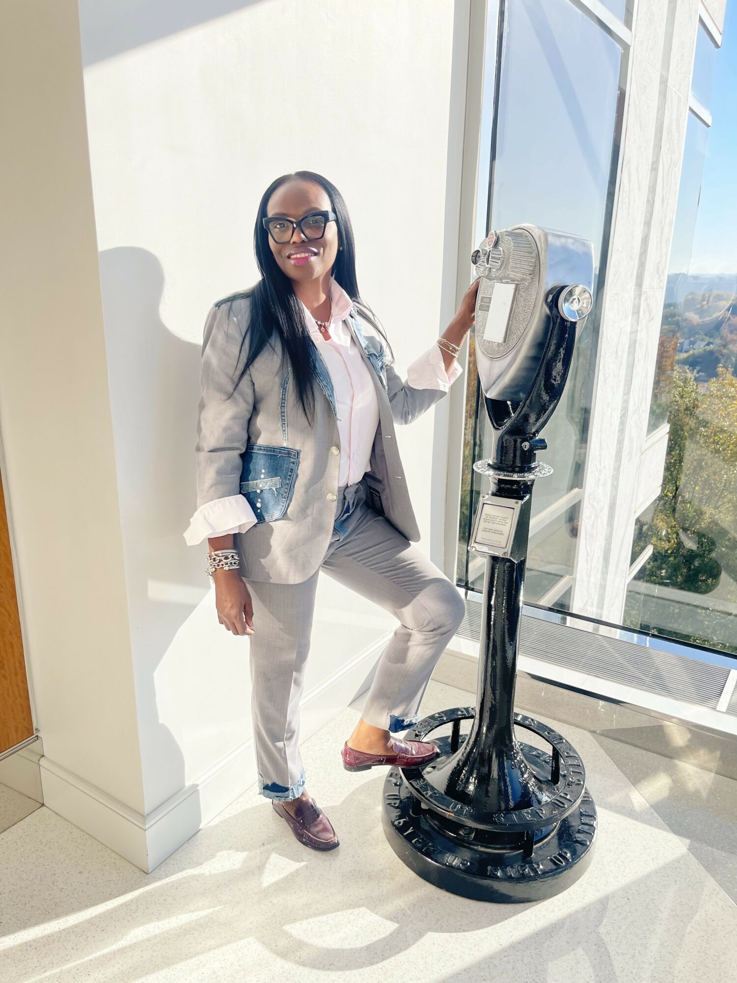 Woman in light gray suit with glasses standing next to a coin-operated viewing telescope, smiling, with large windows and a view outside.