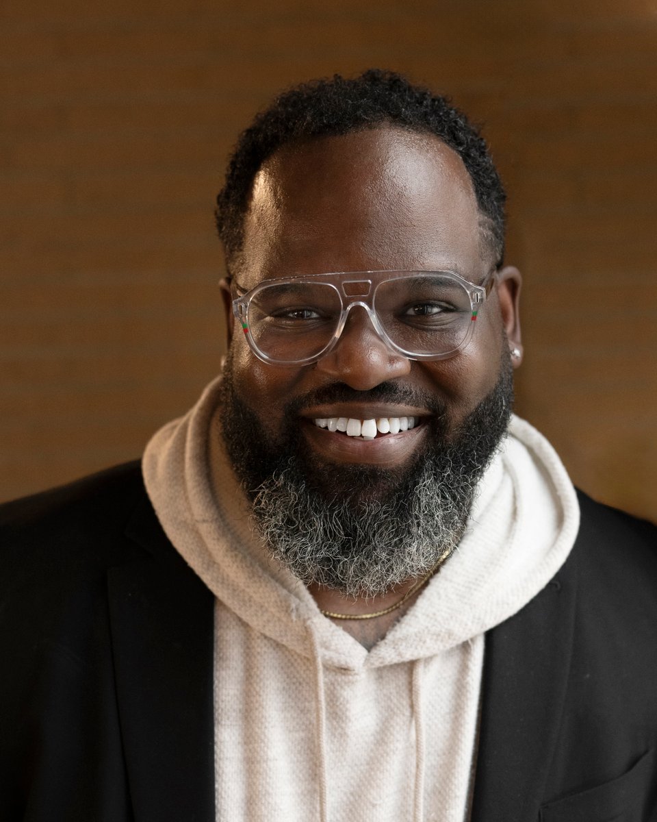Smiling man with glasses, beard, and short curly hair, wearing a hoodie and jacket, against a brown background.