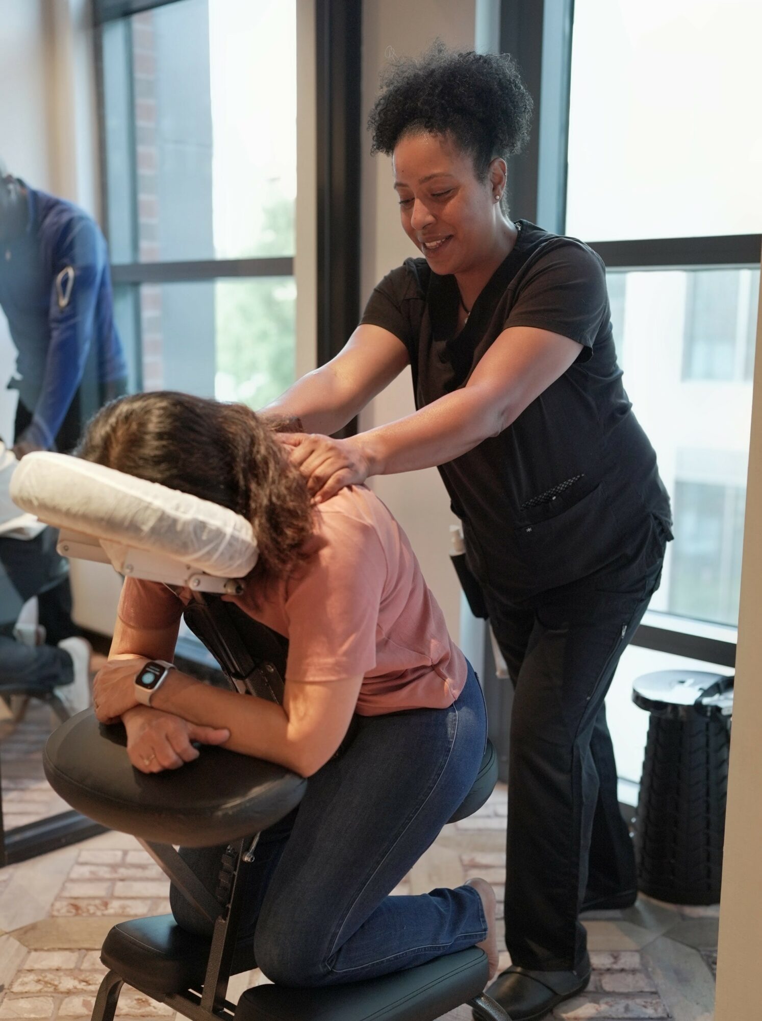 Person receiving a massage or therapy from a smiling woman in a black uniform, seated in a chair near large windows.