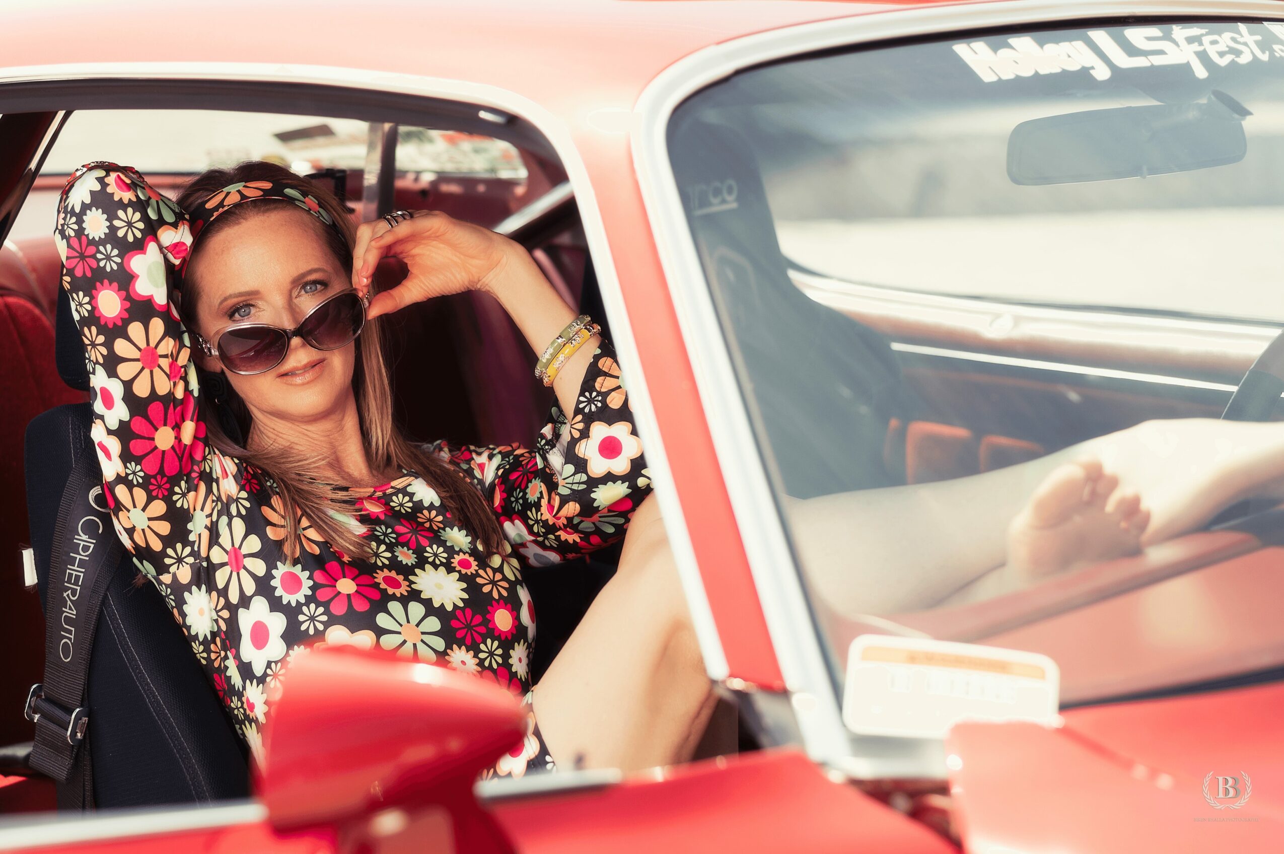 Woman with sunglasses sitting in a car, wearing a floral patterned top, with her arm resting on the window frame.